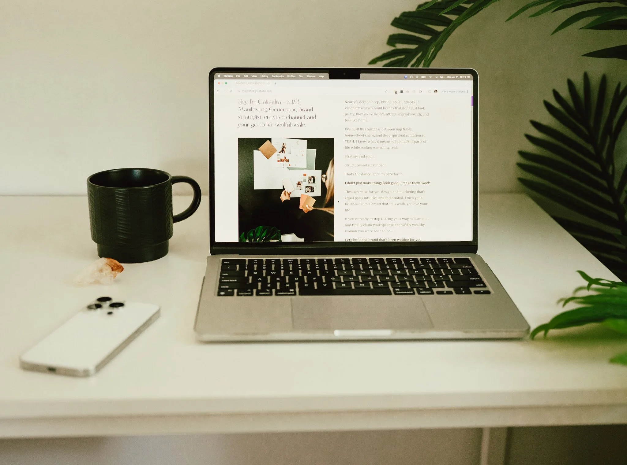 A MacBook laptop on a white desk, displaying a website with text and images. To the left of the laptop is a black mug and some small black objects, possibly pills. There is a green plant on the right side of the image and a rock or mineral in the bottom left corner. The background is a plain, light-colored wall.