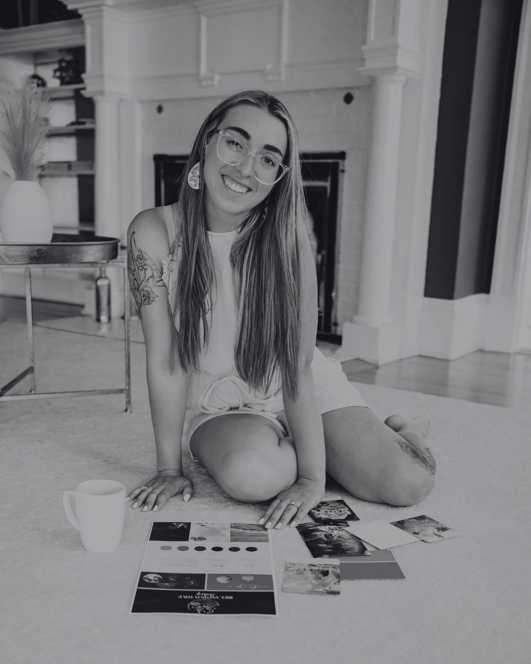 A young woman with long hair, glasses, and tattoos is sitting on the floor, smiling at the camera, with photos, color swatches, and a mug in front of her in a living room with a fireplace and decorative elements.