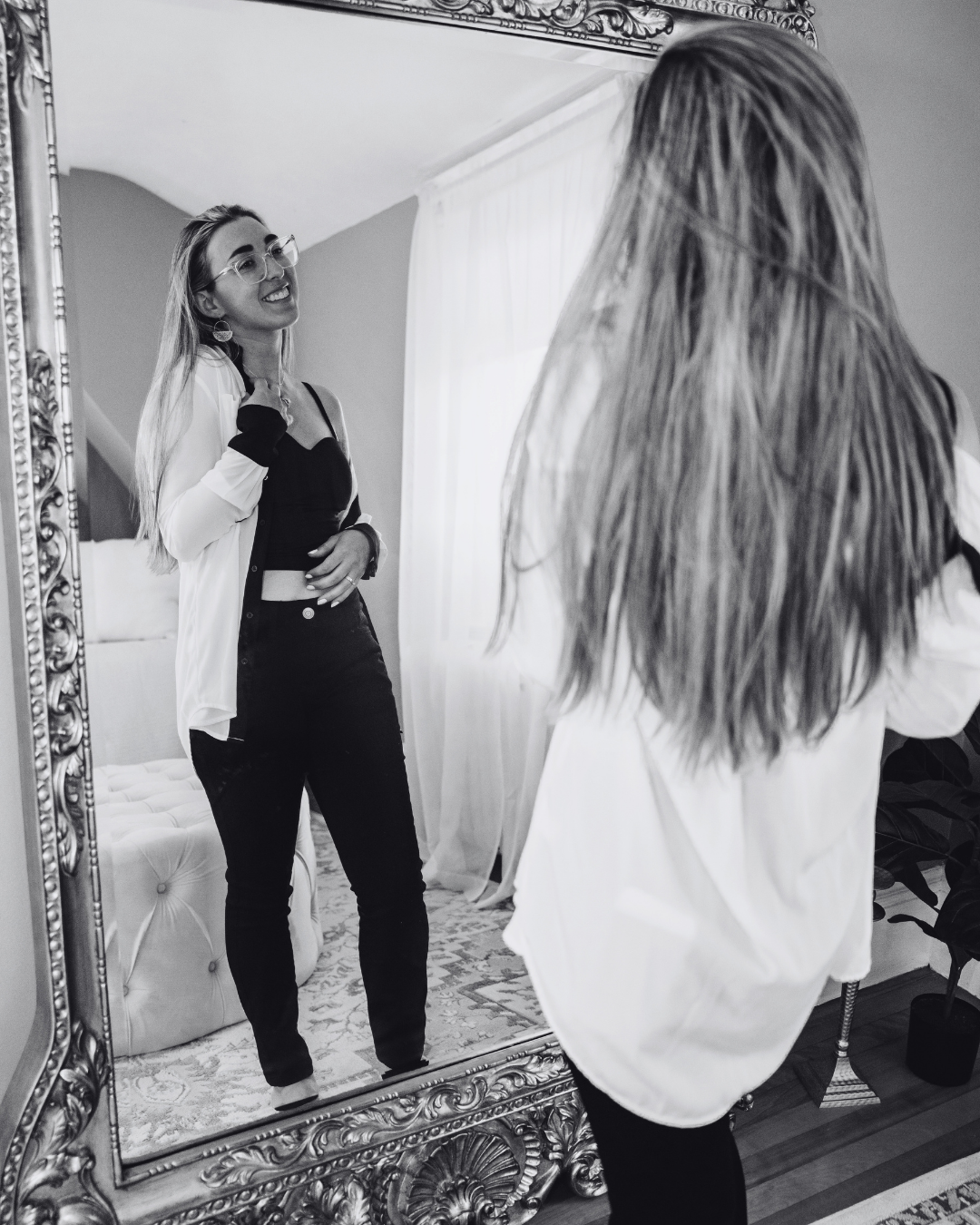 A young woman with glasses, long hair, and a white jacket is smiling in front of a large ornate mirror, adjusting her jacket. Her reflection reveals her black outfit and accessories, and she appears to be in a well-decorated room with natural light coming through a window.
