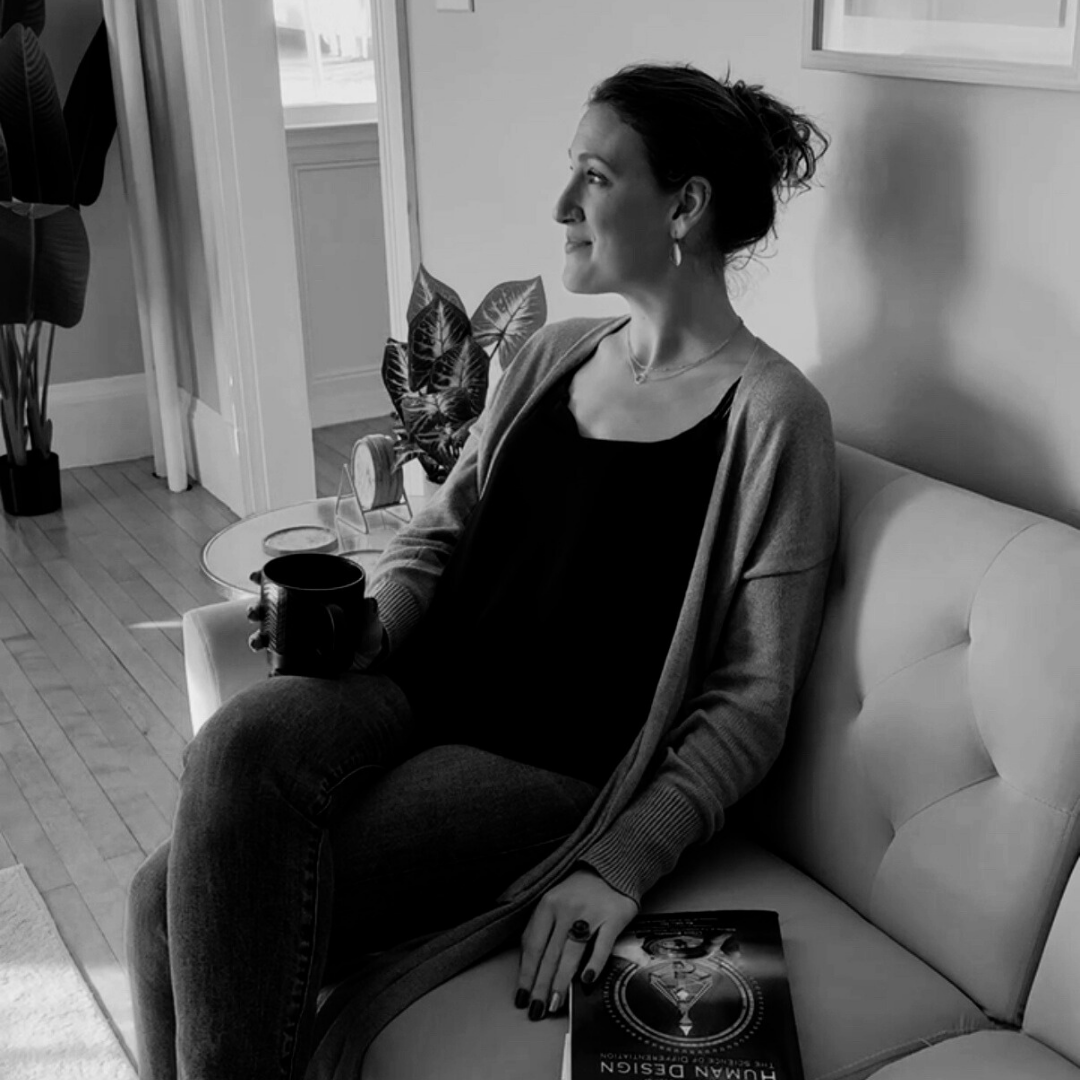 A woman sitting on a white bench or sofa, holding a cup, with a magazine titled 'HUMAN DESIGN' on her lap, in a room with a wooden floor, a large potted plant, and a small side table with an alarm clock, a plant, and a picture frame.
