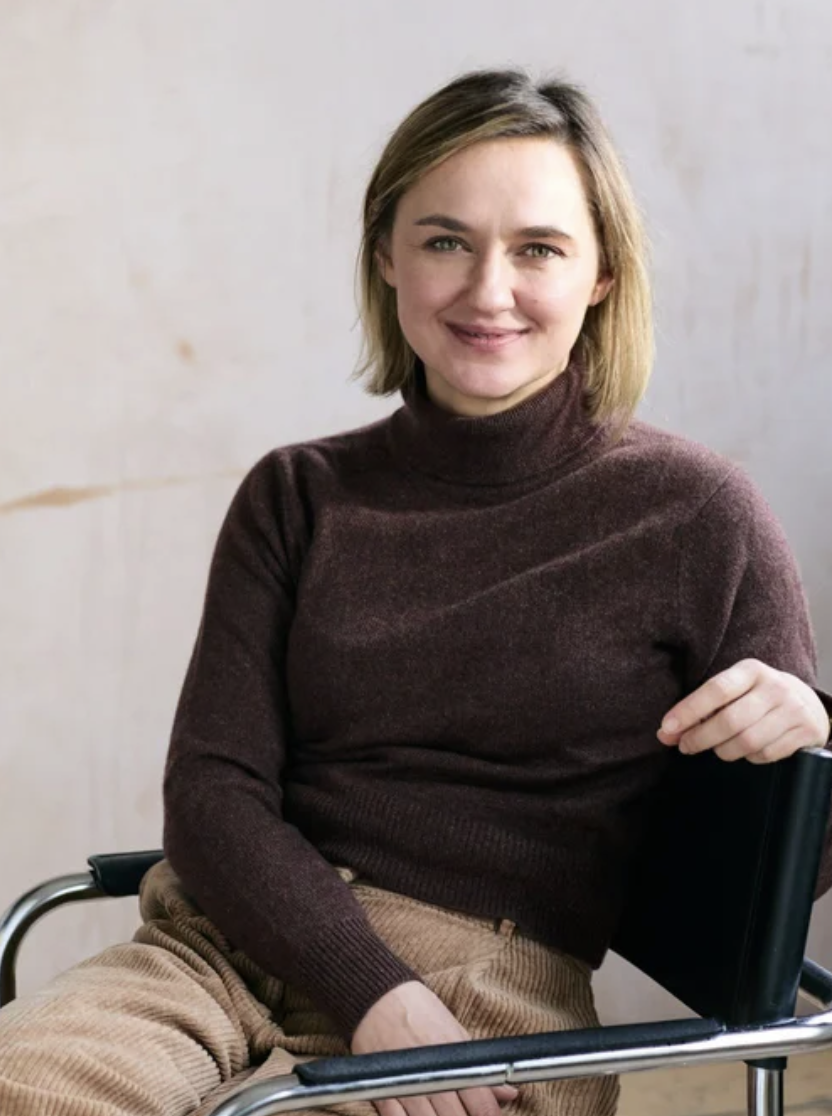 A woman with short blonde hair smiles at the camera while sitting in a chair, wearing a dark brown turtleneck jumper