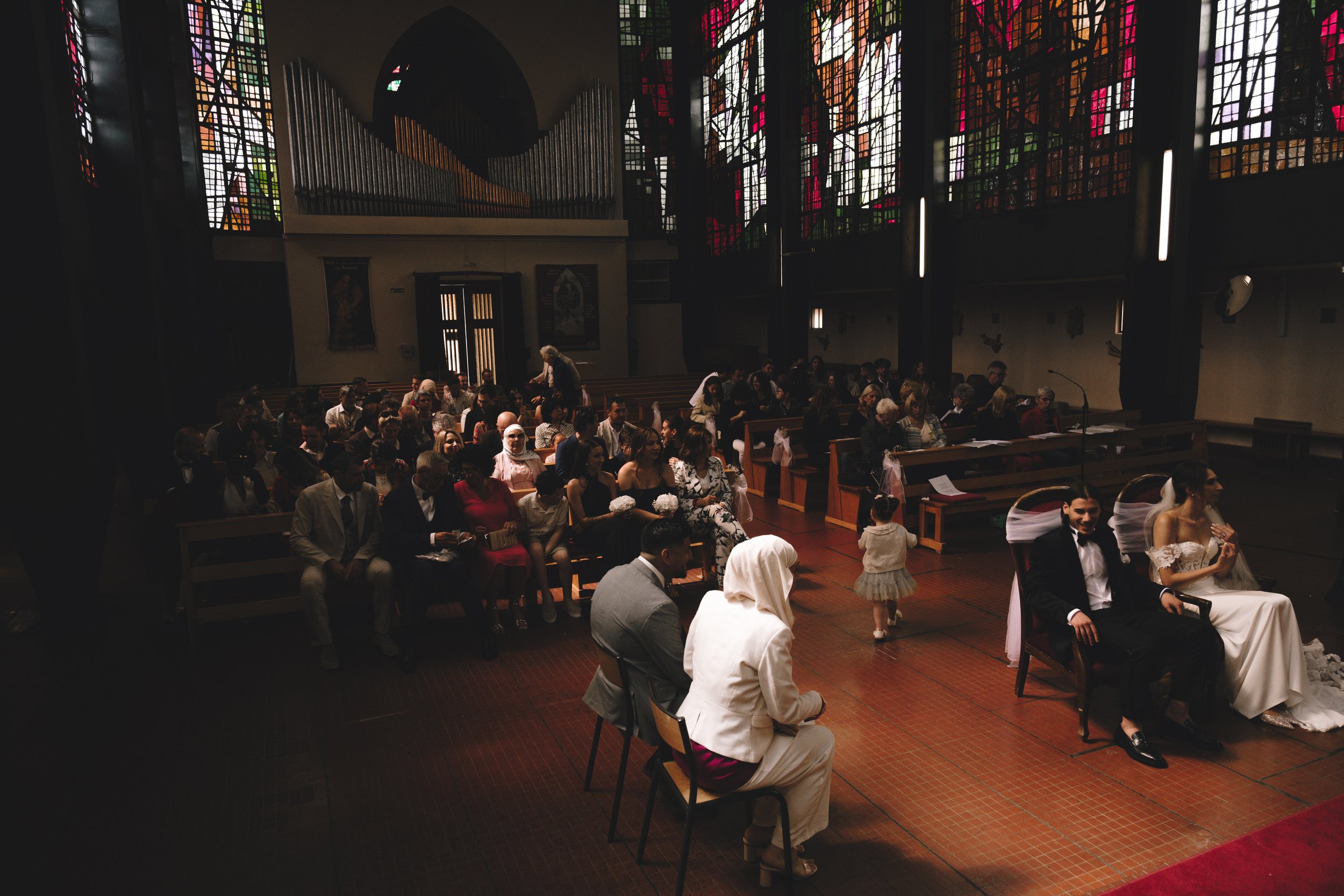 Cérémonie de mariage dans une église avec des invités assis, un couple de mariés devant, et des vitraux en arrière-plan.