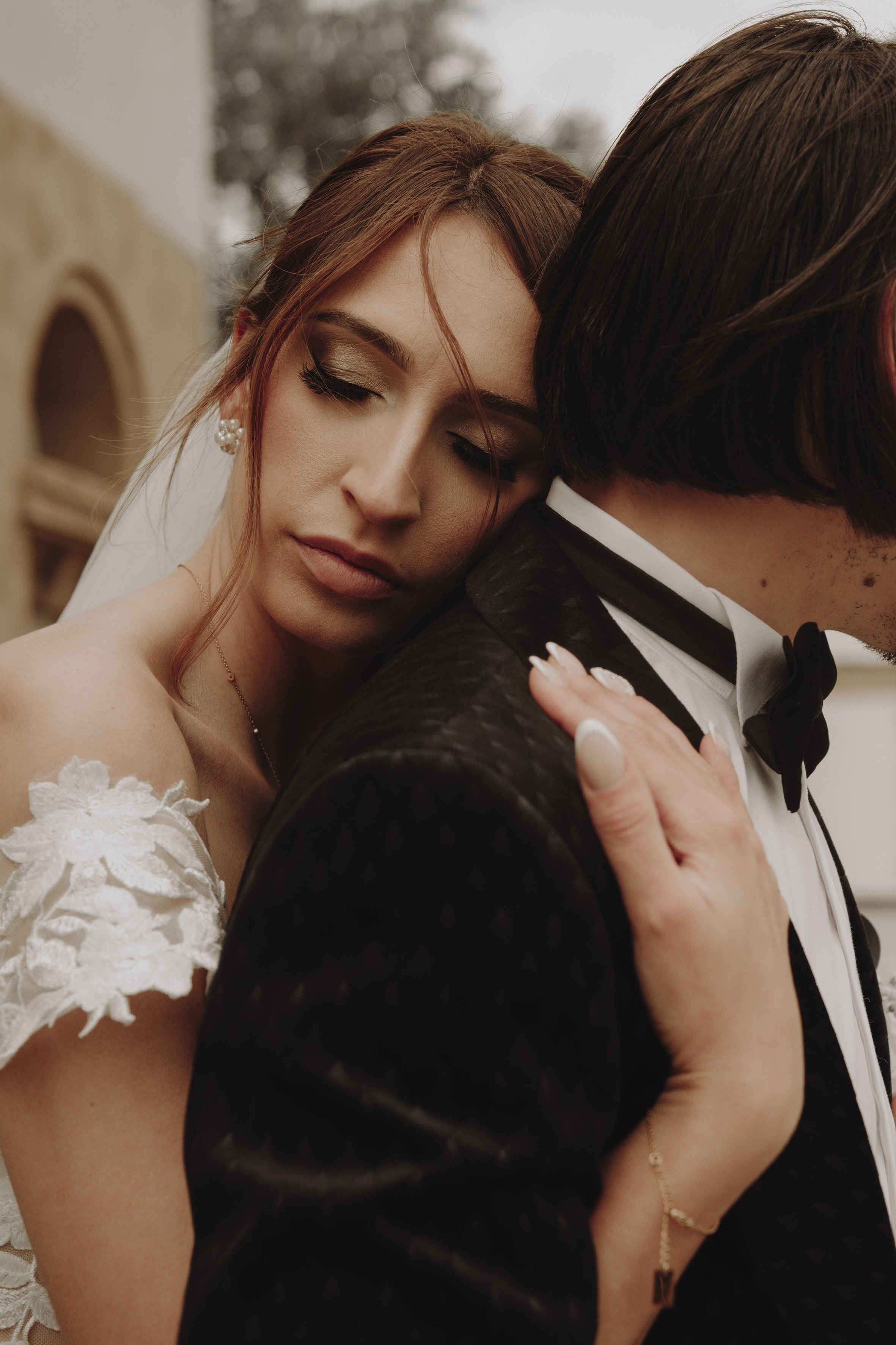 Une mariée en robe blanche avec une épaule en dentelle repose sa tête sur l'épaule d'un homme en smoking noir.