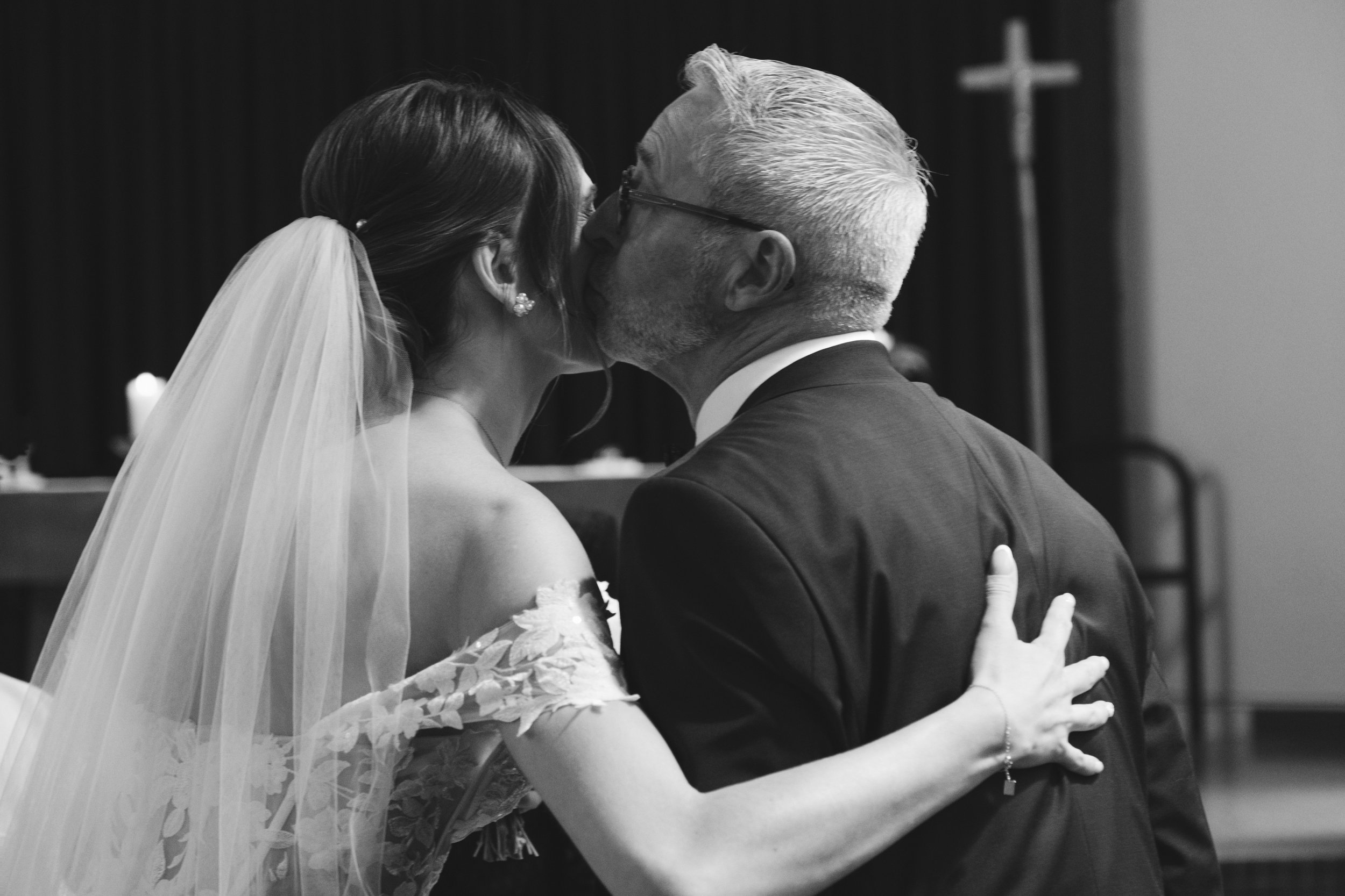 Une mariée embrasse un homme plus âgé, possiblement son père, lors d'une cérémonie. Ils sont en tenue formelle, la mariée porte un voile et une robe en dentelle.