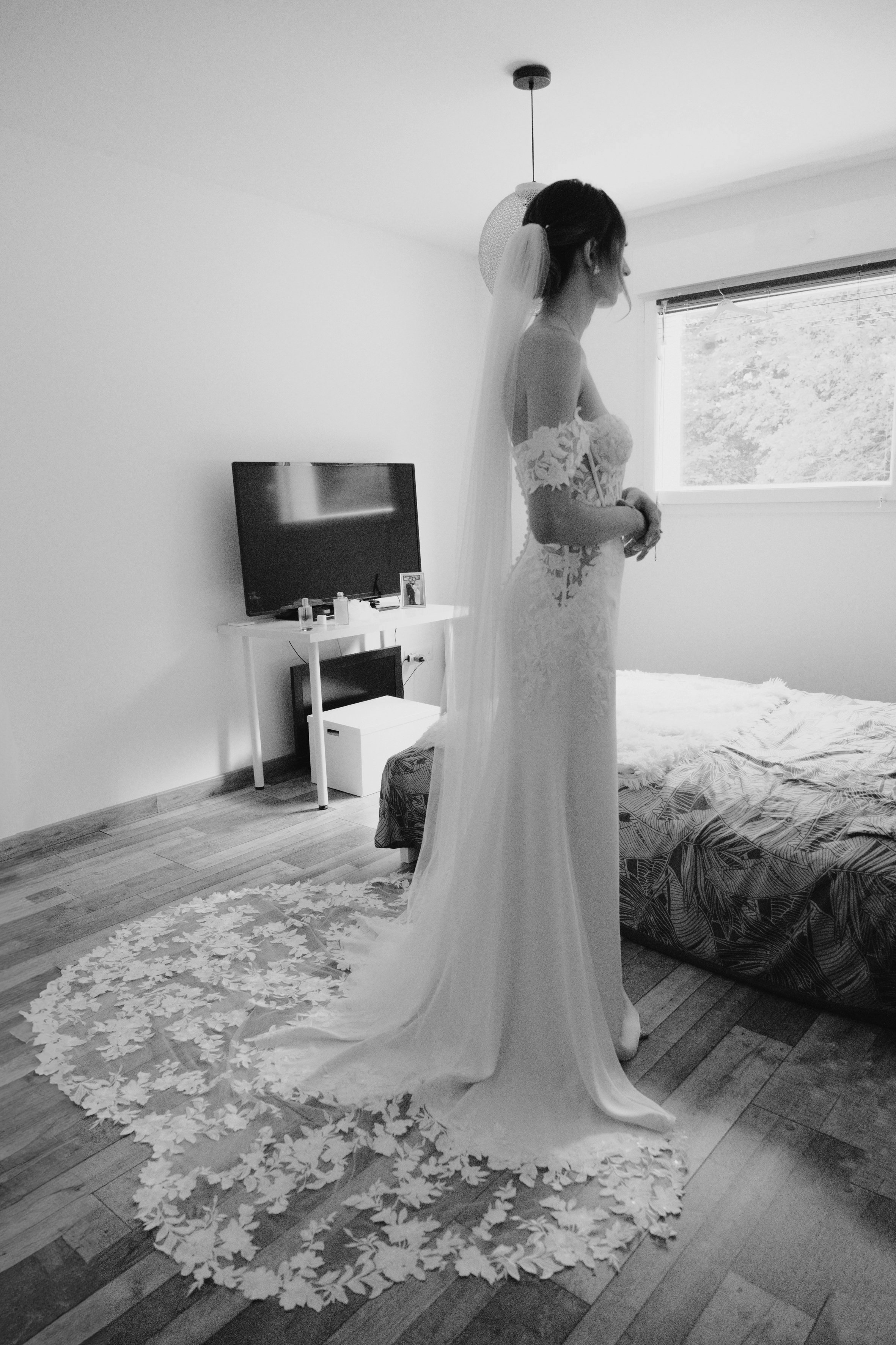 Une femme en robe de mariée se tient dans une chambre, devant un lit et un bureau avec une télévision.
