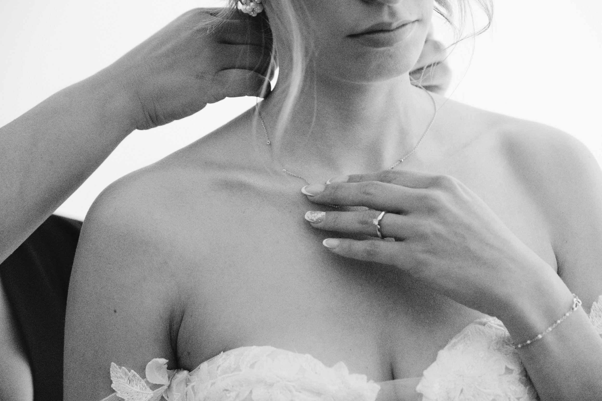 Femme portant une robe de mariée, ajustant un collier, avec une main tenant le fermoir dans le dos, vue en noir et blanc.