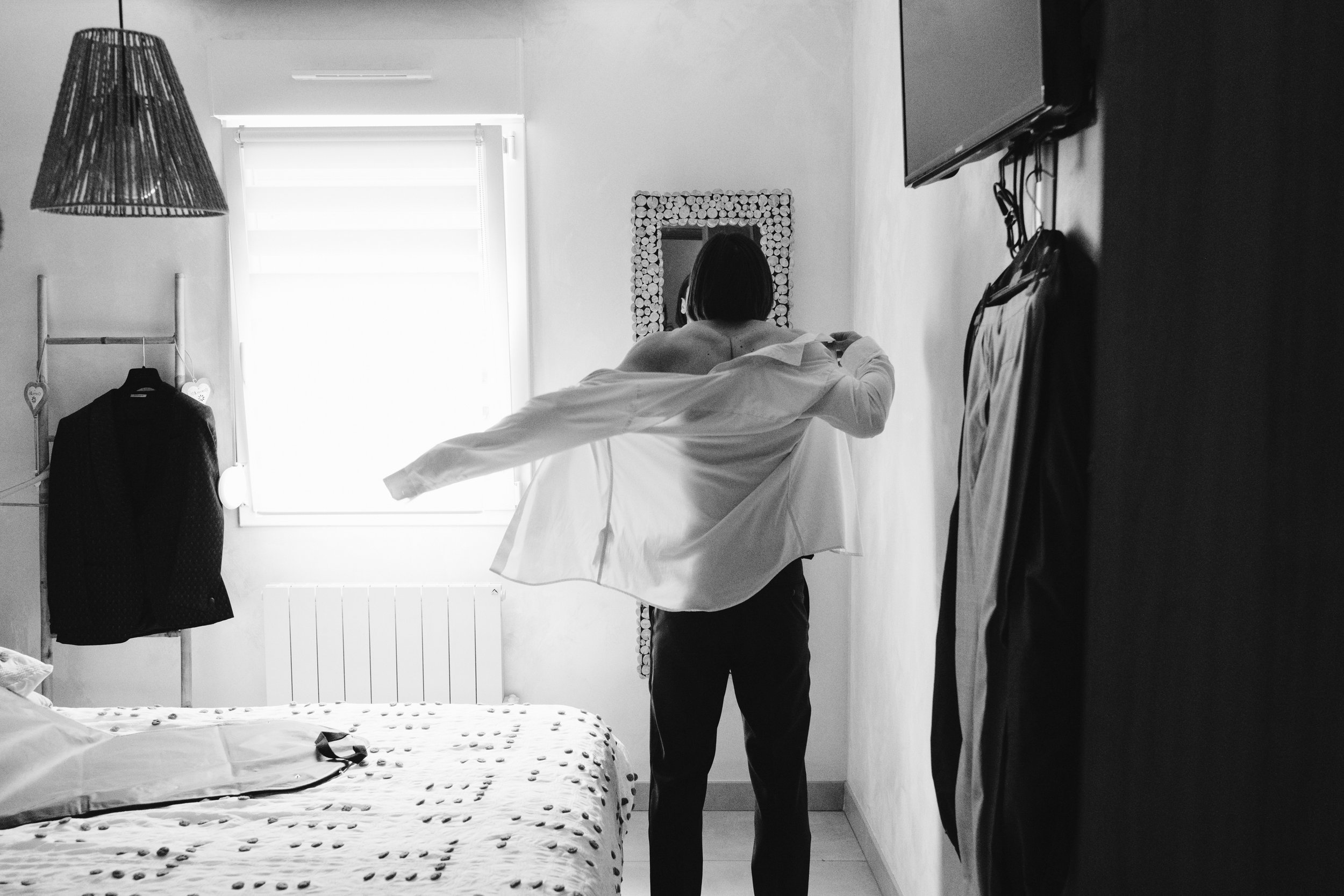 Personne en train de mettre une chemise dans une chambre, vêtements accrochés près d'eux, lampe suspendue, couvre-lit à motifs, fenêtre derrière.