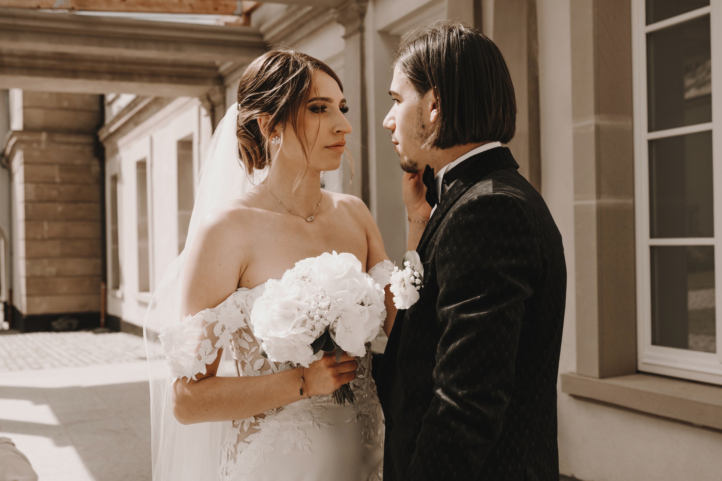 Couple de mariés posant ensemble, la mariée en robe blanche tenant un bouquet de fleurs, le marié en costume sombre, dehors près d'un bâtiment.