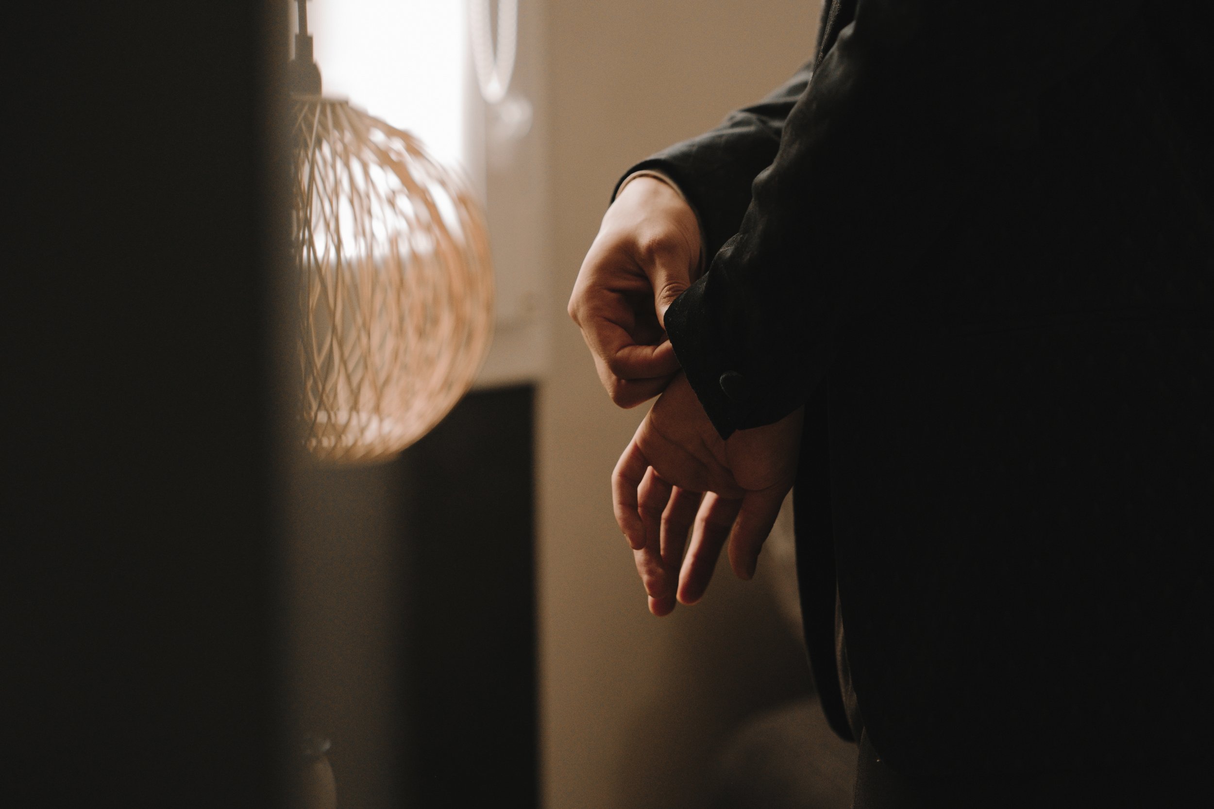 Une personne ajuste un bouton de manchette d'une veste sombre, près d'un abat-jour en rotin.