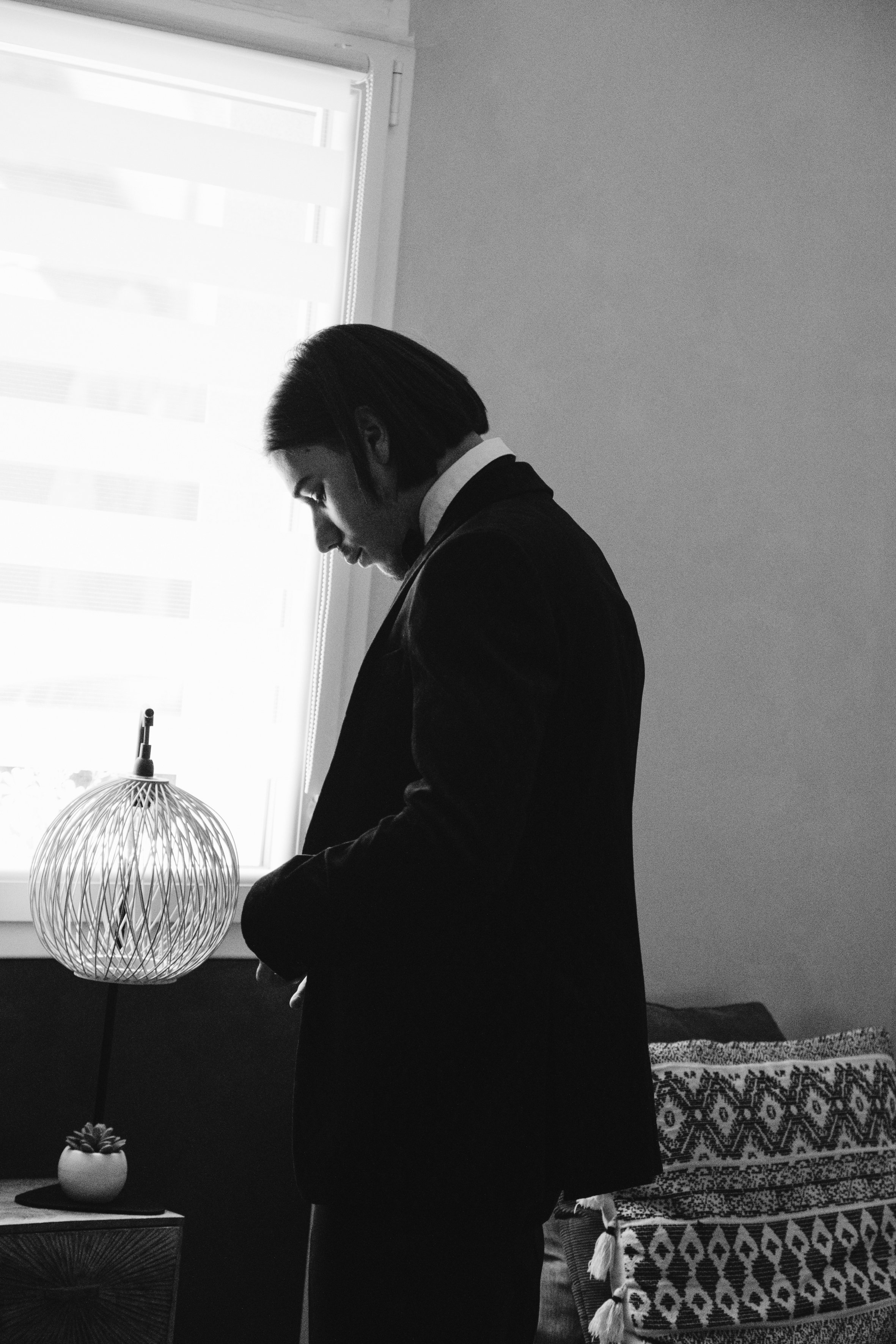 Homme en costume debout près d'une fenêtre, lampe moderne sur une table, ambiance monochrome.