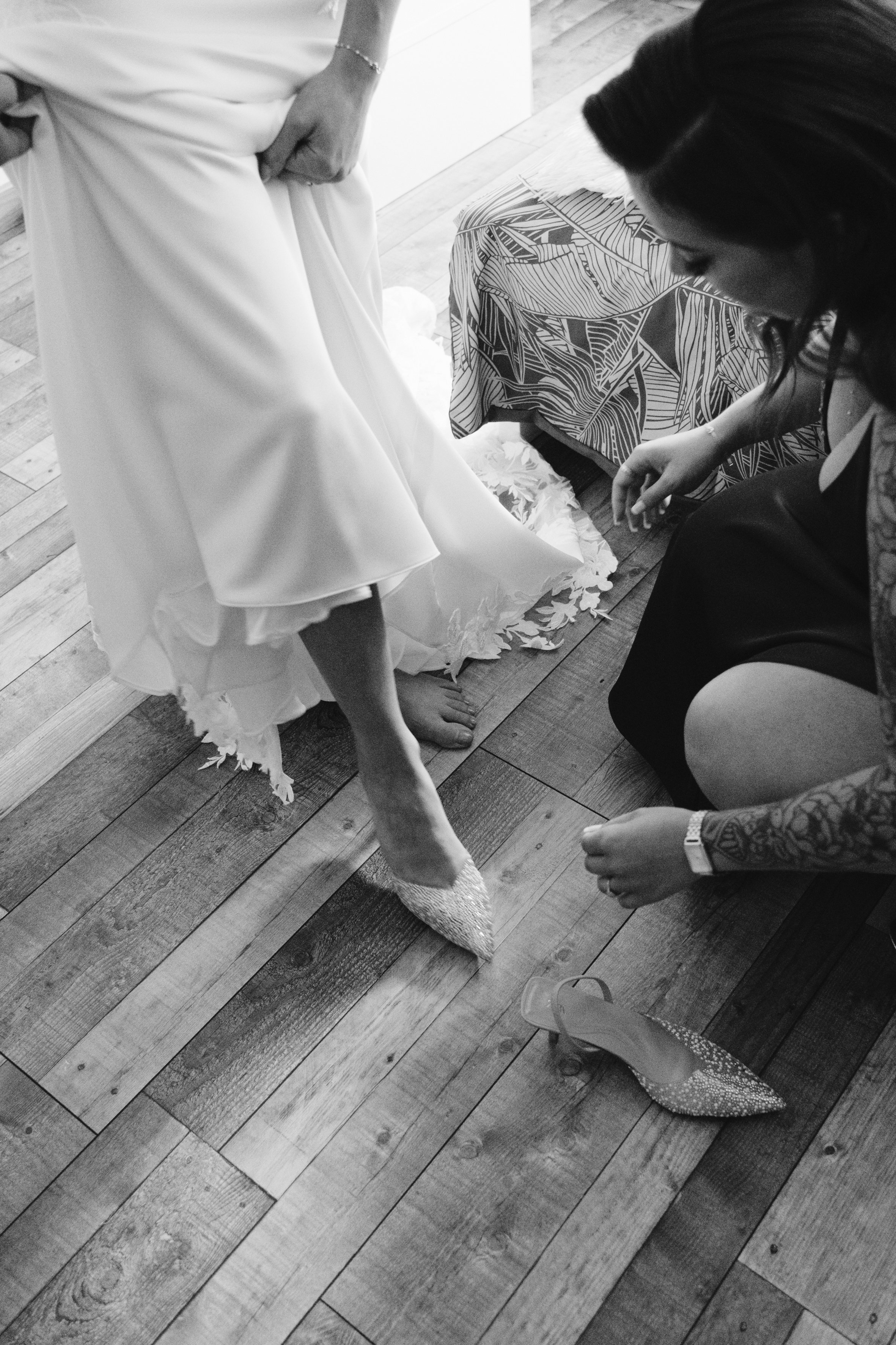 Personne portant une robe blanche enfilant des chaussures élégantes avec l'aide d'une autre personne, sur un sol en bois.