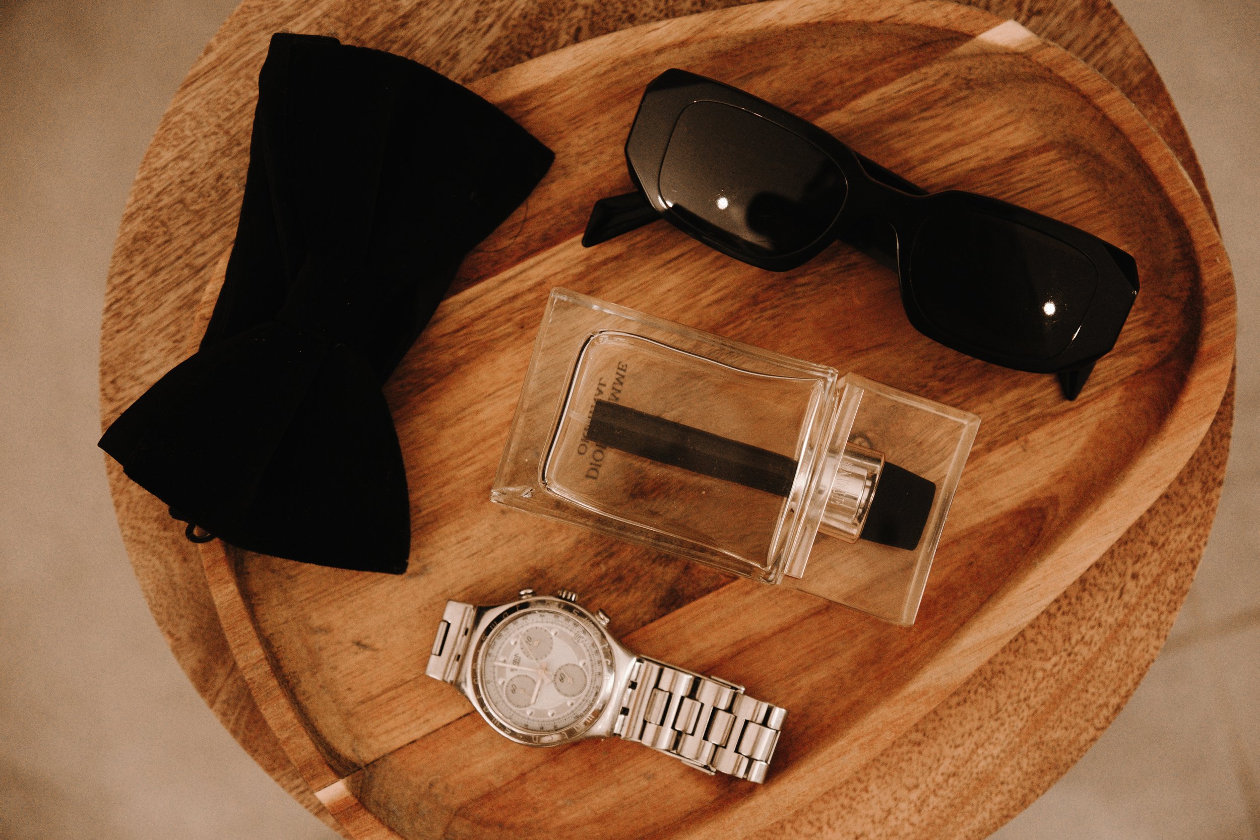 Montre en métal, lunettes de soleil noires, flacon de parfum, et nœud papillon noir sur une table en bois.