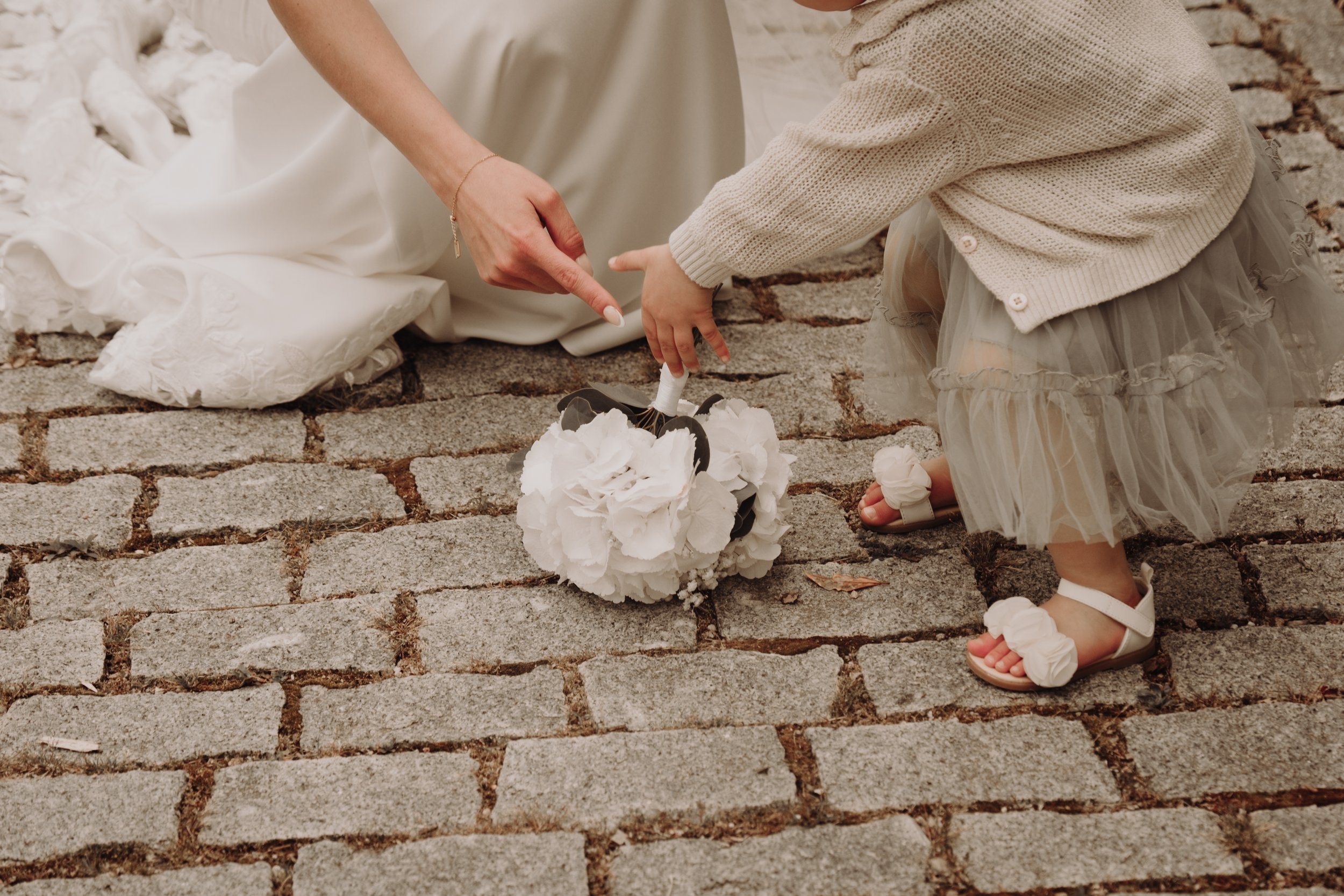 Une femme en robe de mariée avec un enfant portant une jupe en tulle et des sandales blanches, touchant un bouquet de fleurs blanches sur un sol pavé.