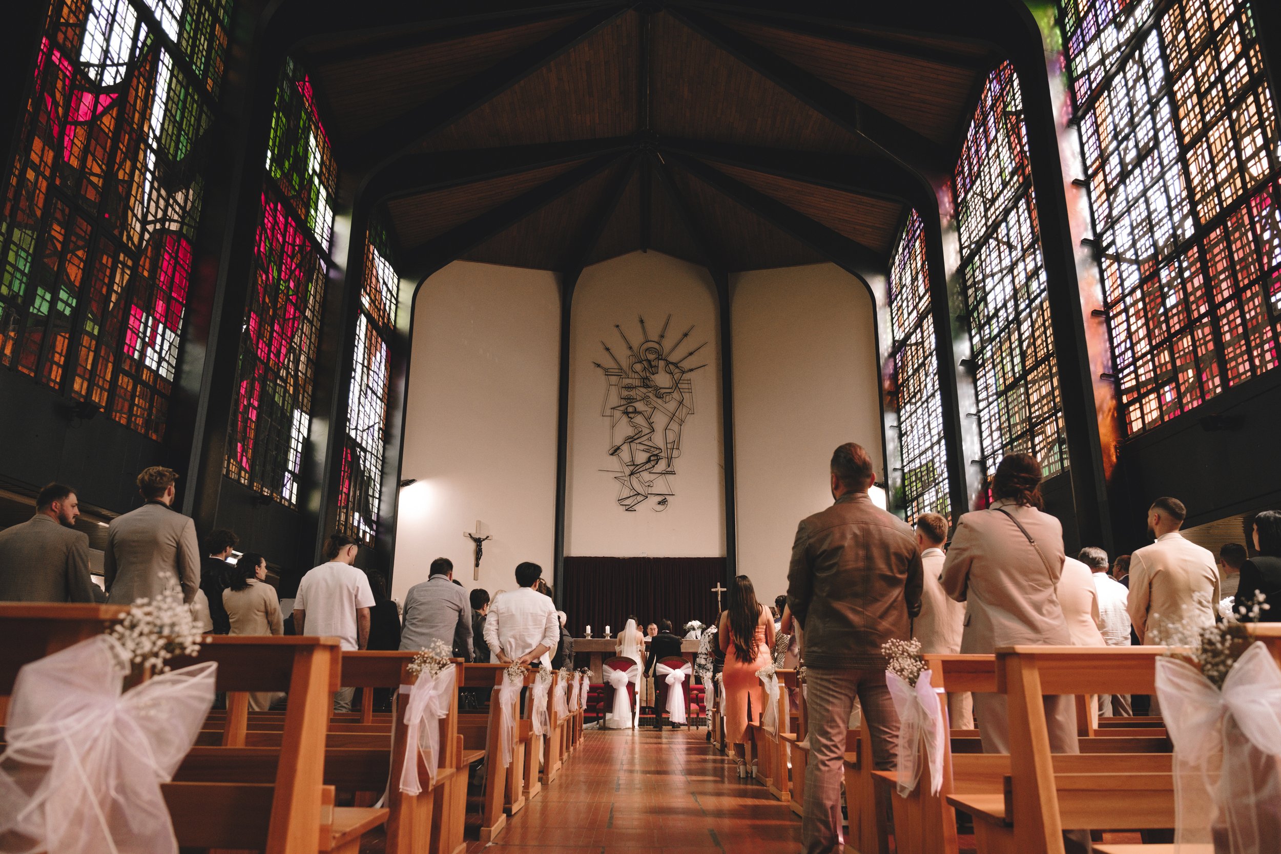 Intérieur d'une église avec des vitraux colorés et des fidèles assis aux bancs. Un couple est devant l'autel, probablement pour une cérémonie de mariage.