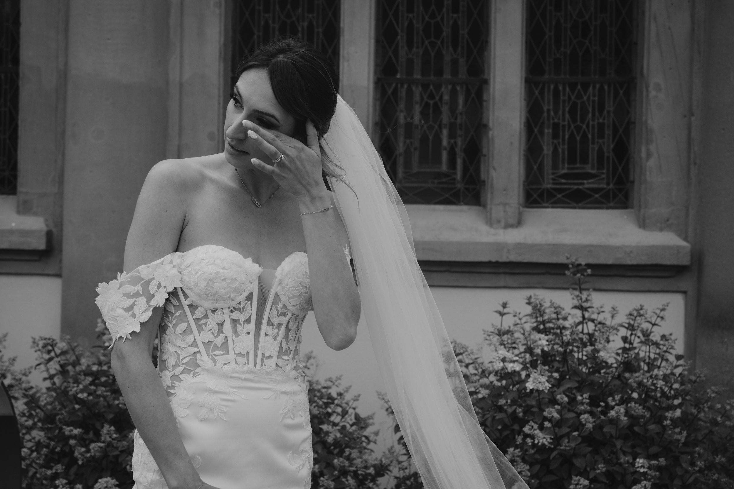 Mariée en robe blanche essuyant ses larmes, devant une fenêtre avec vitraux, avec des buissons en arrière-plan.