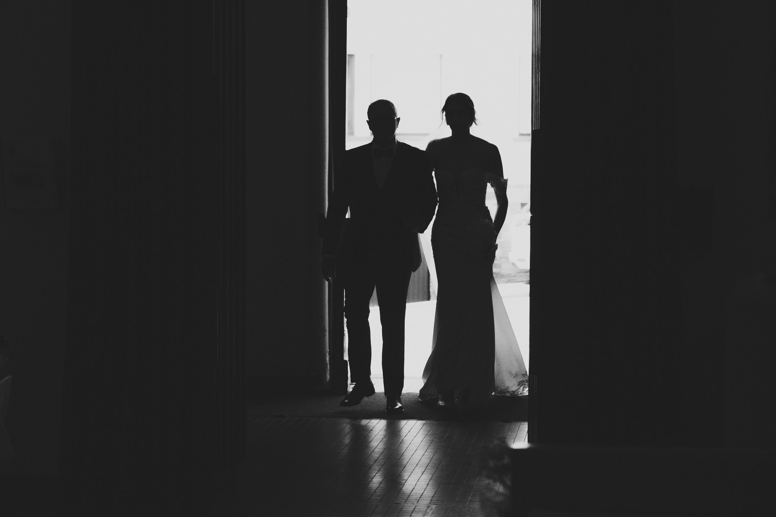 Silhouette d'un couple entrant dans un lieu éclairé par l'arrière, vêtus de tenues formelles de mariage.