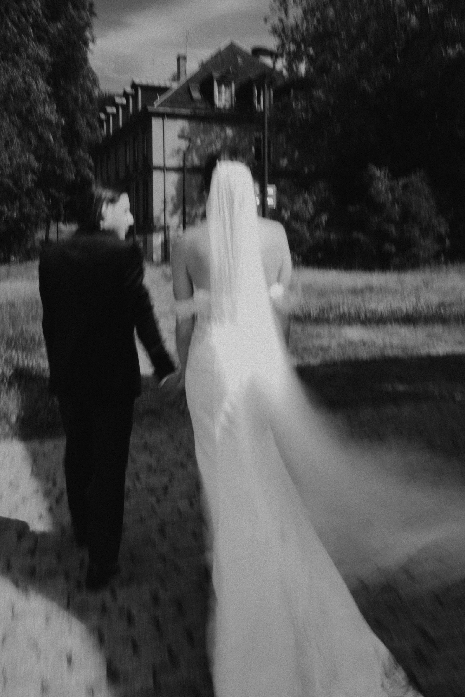 Un couple de mariés marchant main dans la main, vu de dos, avec des arbres et une maison en arrière-plan.