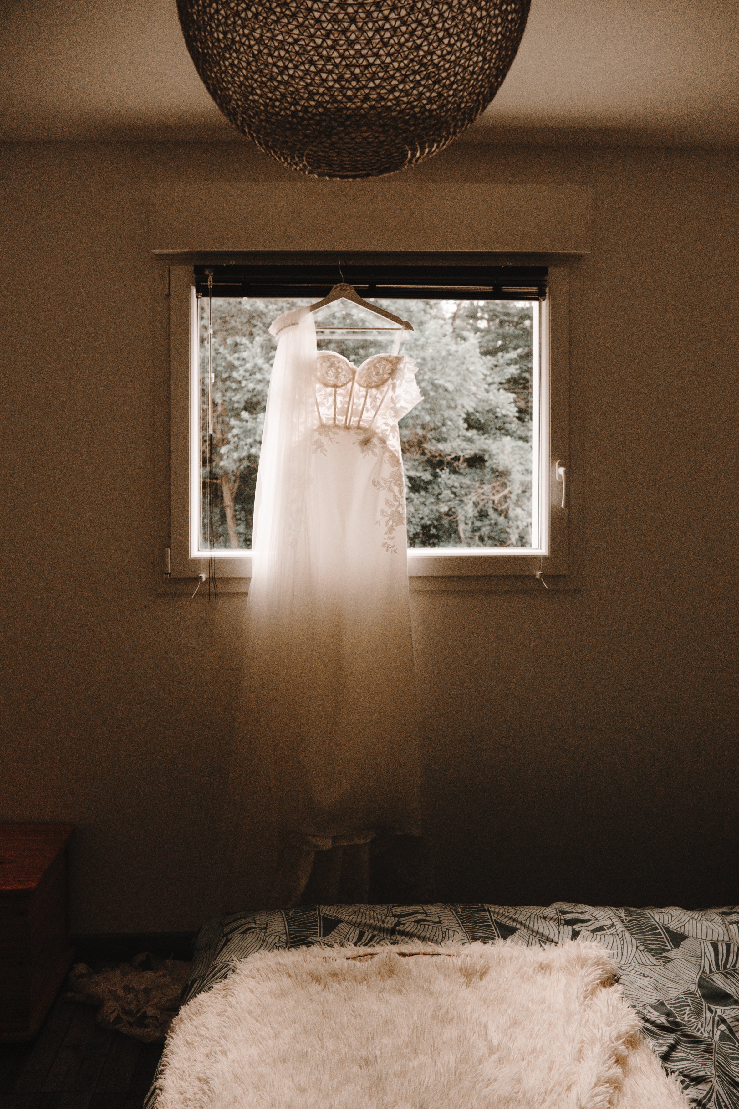 Robe de mariée suspendue devant une fenêtre avec un fond de nature verdoyante, dans une chambre peu éclairée.