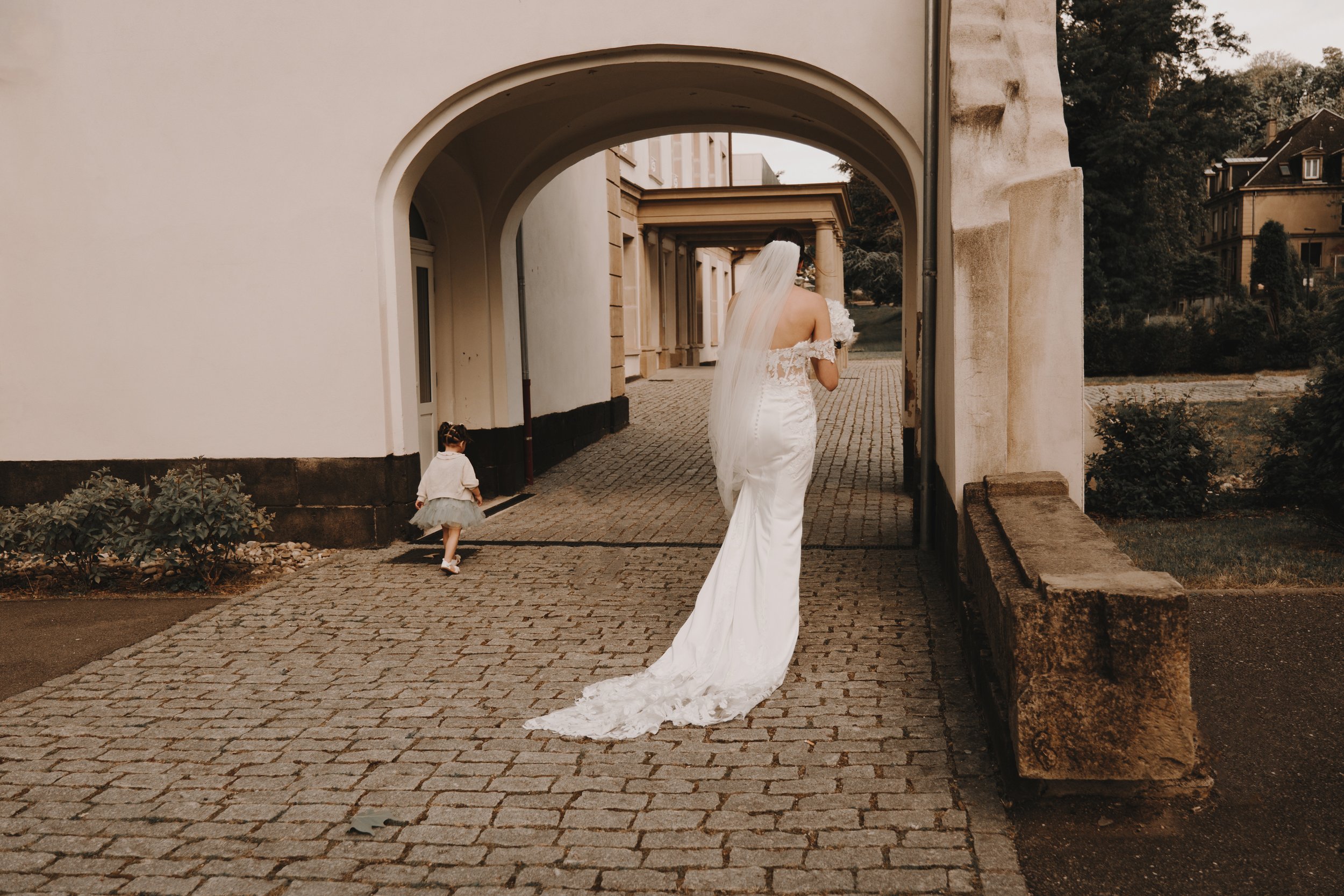 Une mariée en robe blanche marchant sous une arche en pierre avec une petite fille en robe tutu marchant à ses côtés.