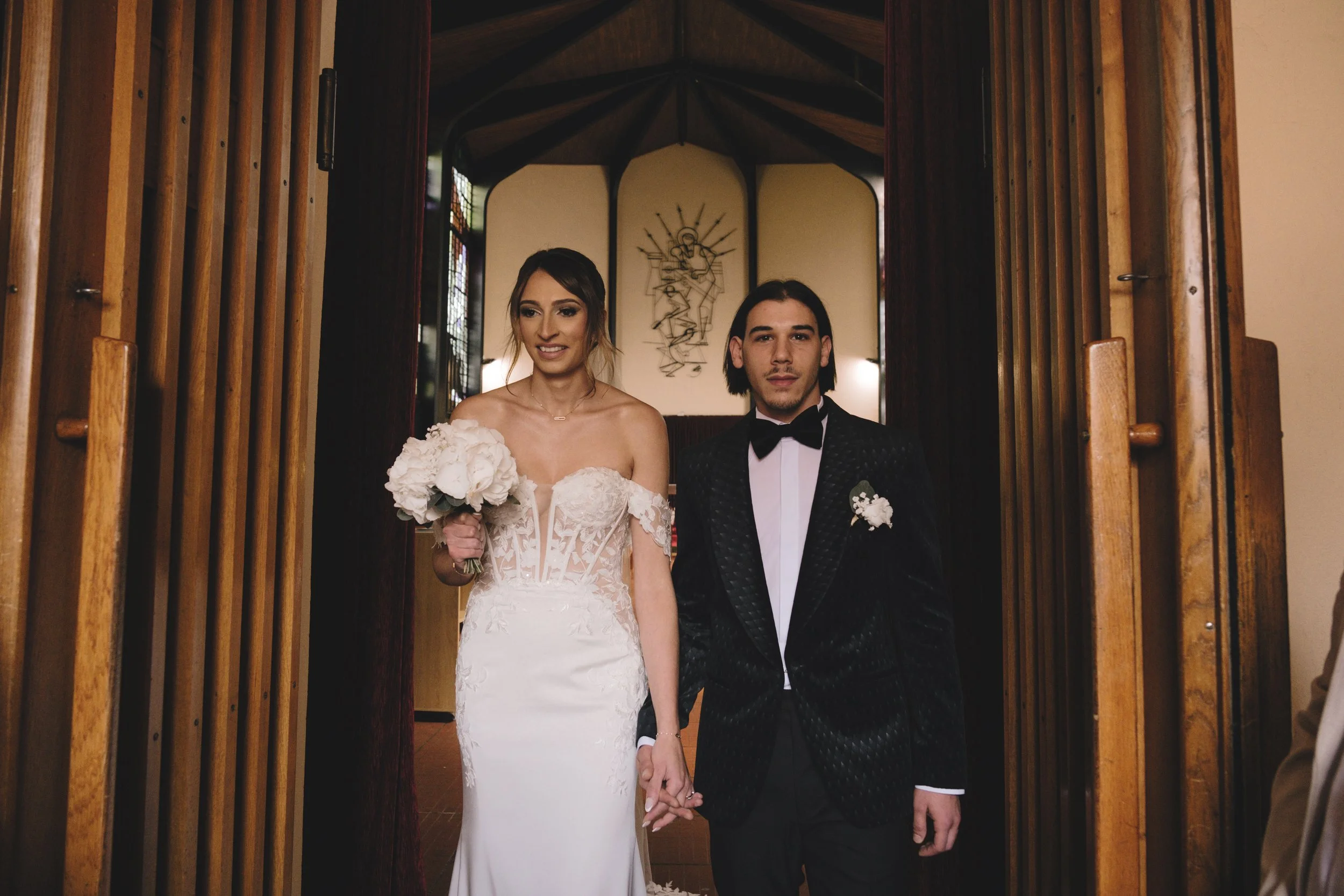 Un couple se tient la main dans un cadre nuptial. La femme porte une robe de mariée blanche et tient un bouquet de fleurs. L'homme porte un smoking avec un nœud papillon. Les deux semblent sortir d'un bâtiment en bois.