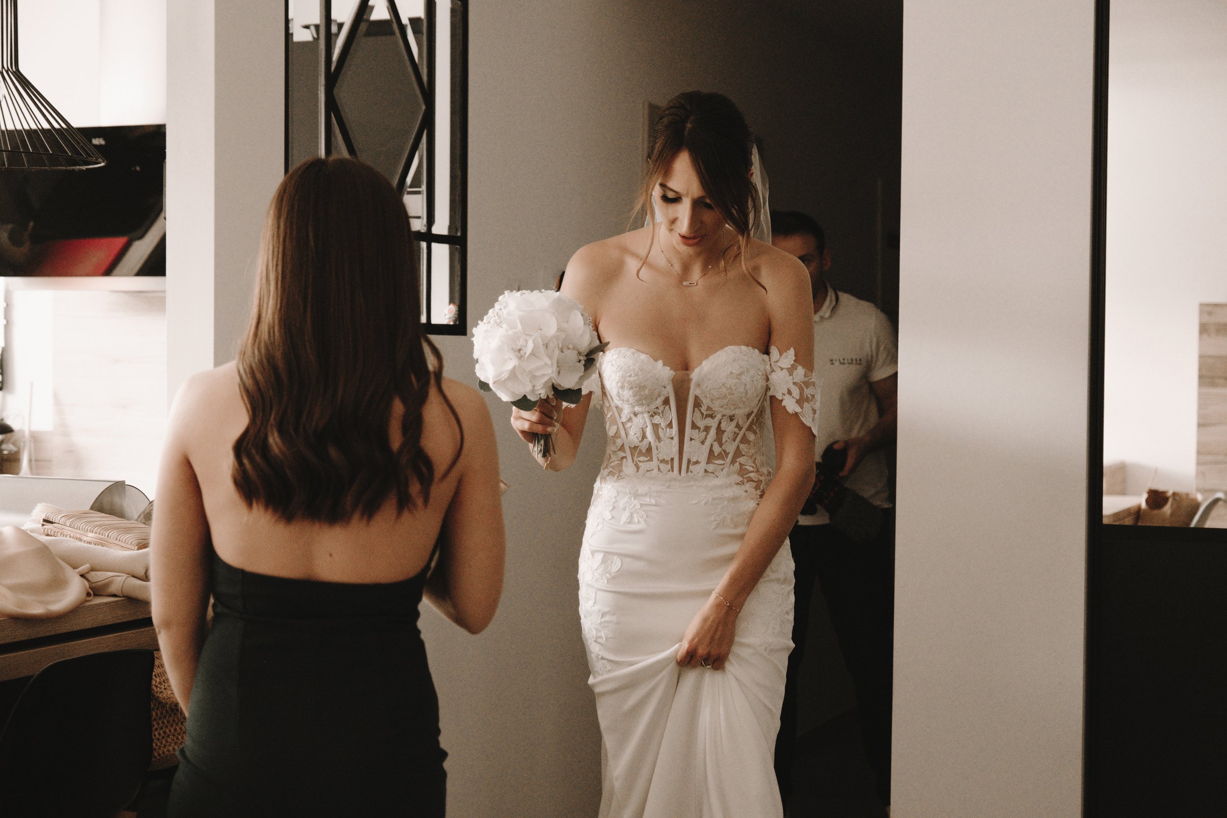 Femme en robe de mariée tenant un bouquet, avec une autre femme en robe noire de dos.