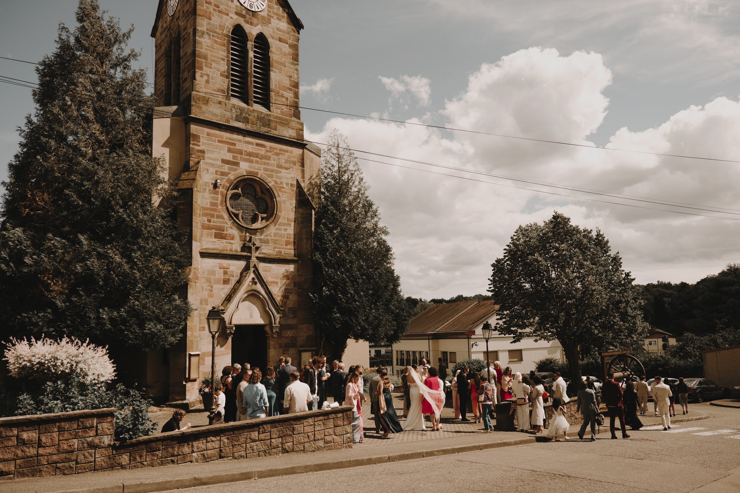 Église en pierre avec groupe de personnes devant, ciel partiellement nuageux.