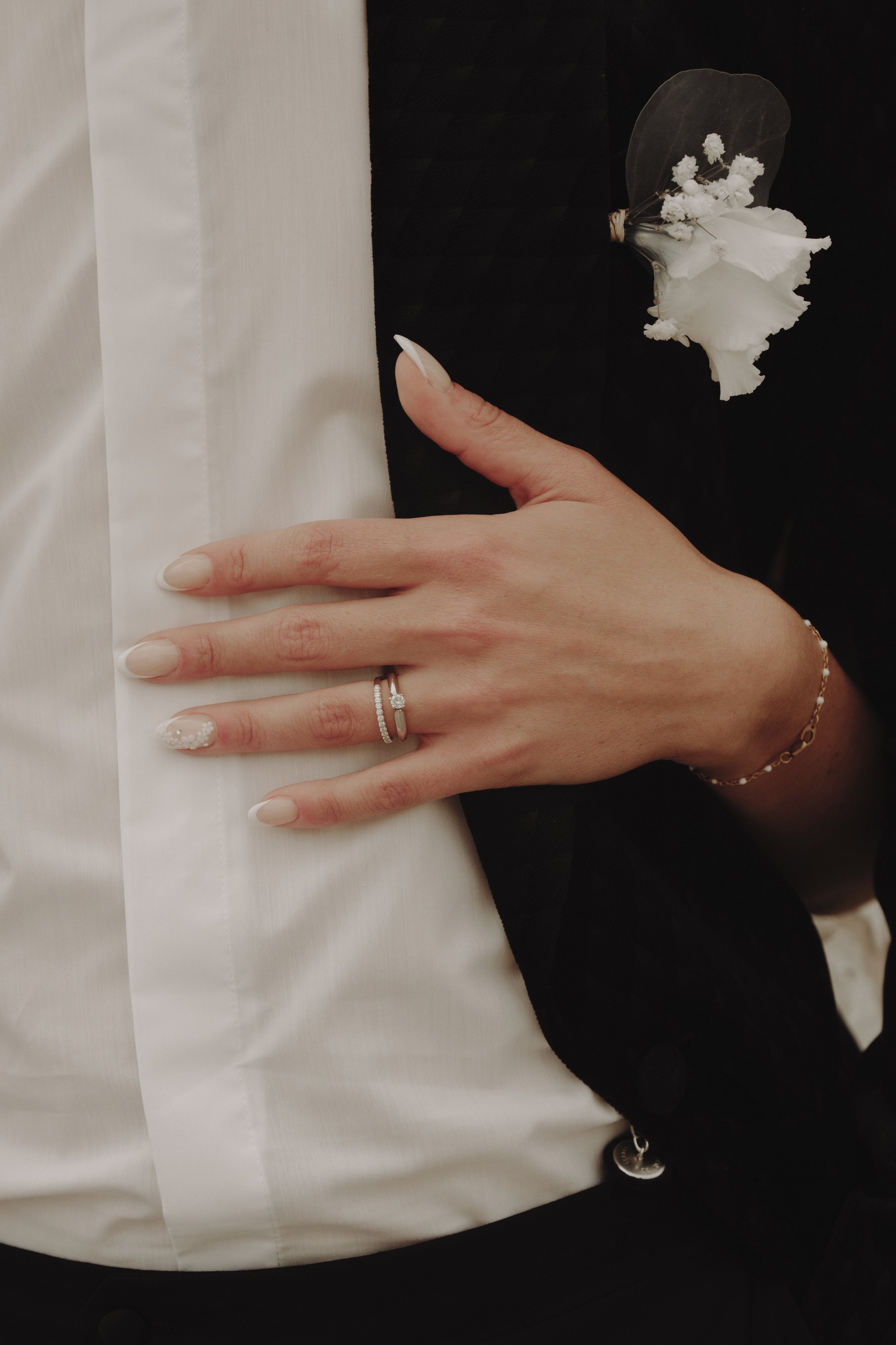Main portant une bague sur une veste noire avec une boutonnière florale blanche.