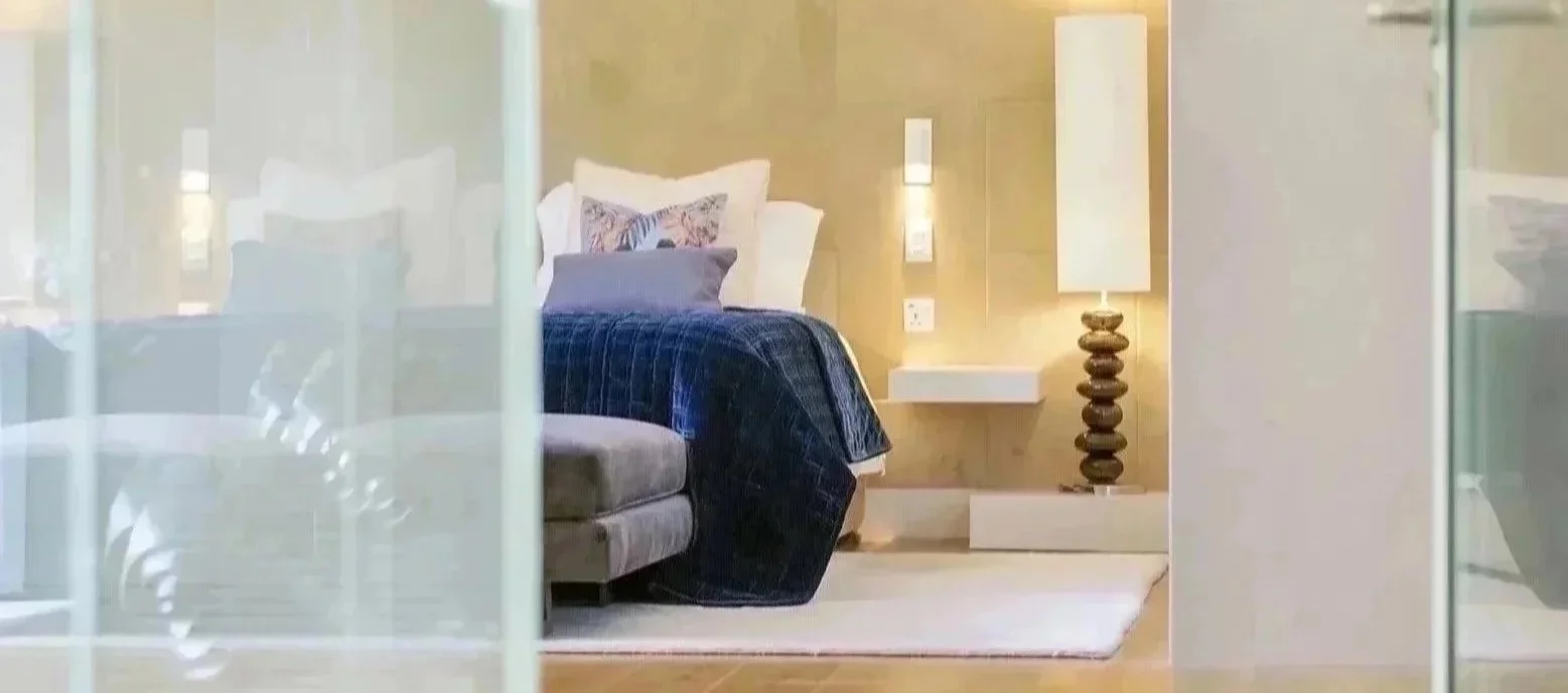 A view of a modern bedroom seen through glass panels, featuring a bed with white and blue bedding, a colorful pillow, a side table with a unique stacked lamp, and a small wall-mounted shelf.