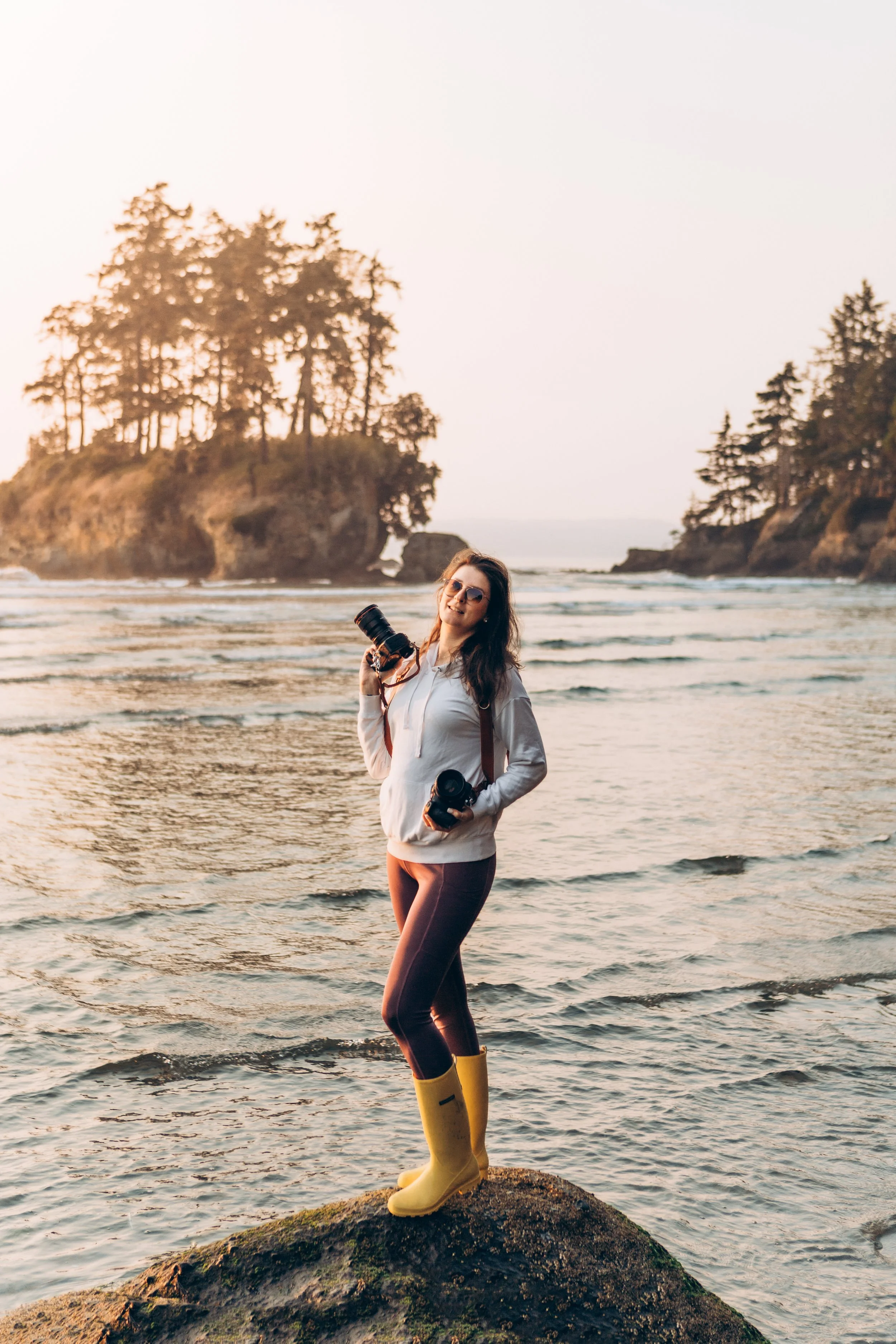 Valerie Lisuk, Pacific Northwest beach elopement photographer, standing on the coast with cameras at sunset