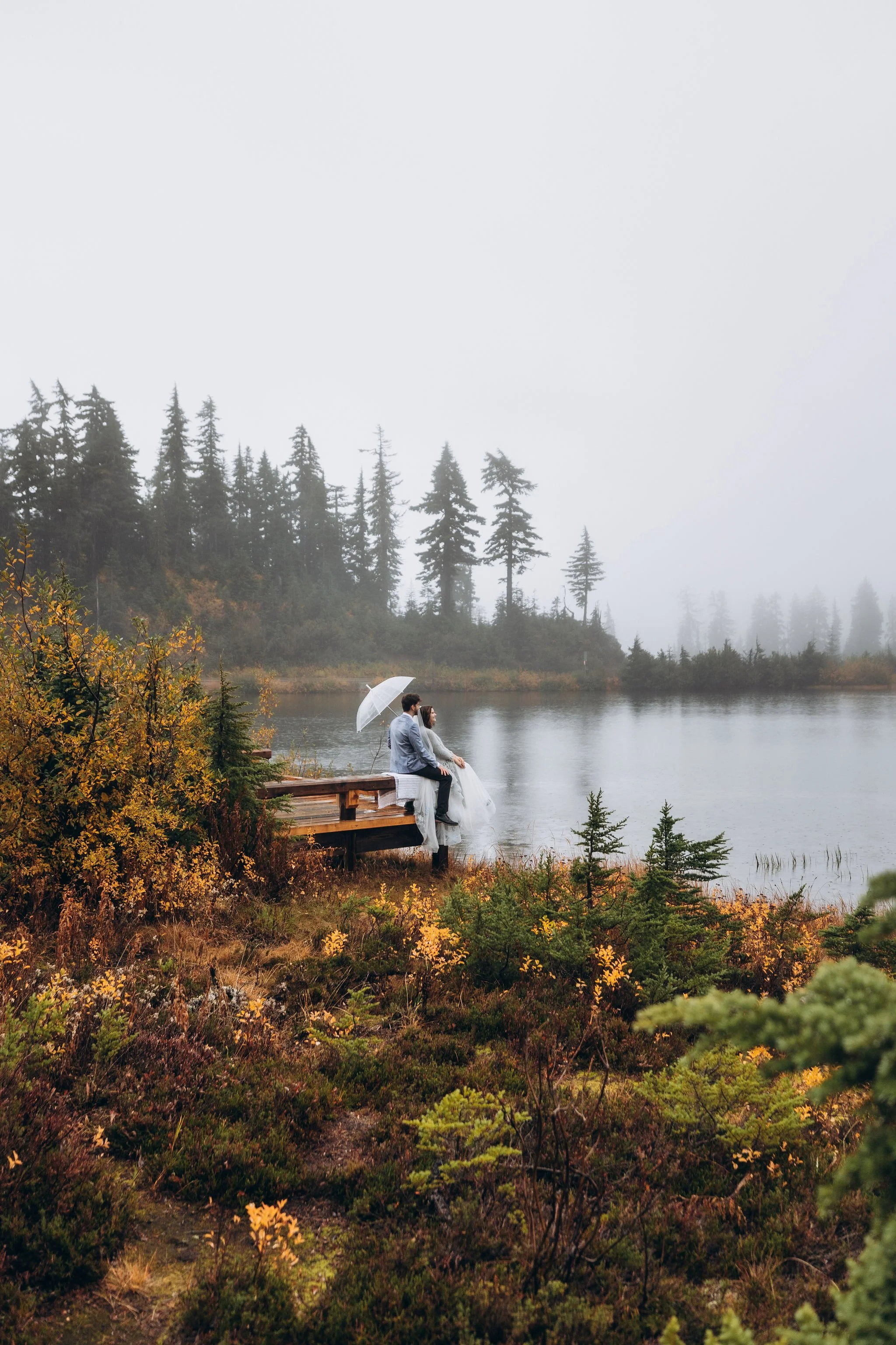 Couple sitting on a wooden dock during a foggy fall North Cascades elopement in Washington with alpine lake and evergreen trees