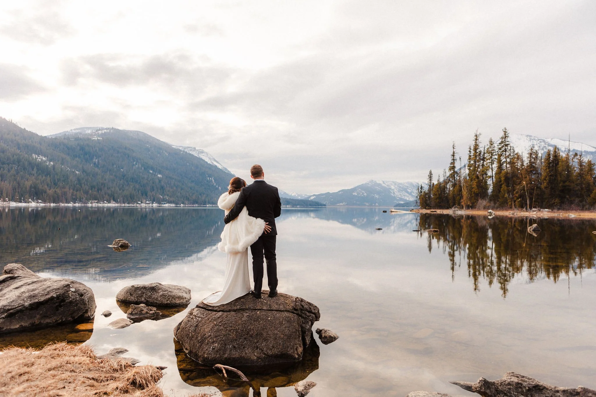 Couple standing on a rock during an elopement at Lake Wenatchee, Washington, with calm water, mountains, and forest reflections