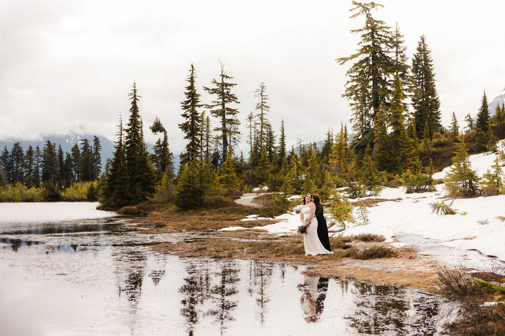 LGBTQ+ couple standing together during a spring elopement at Picture Lake with melting snow, evergreen trees, and reflections in the water