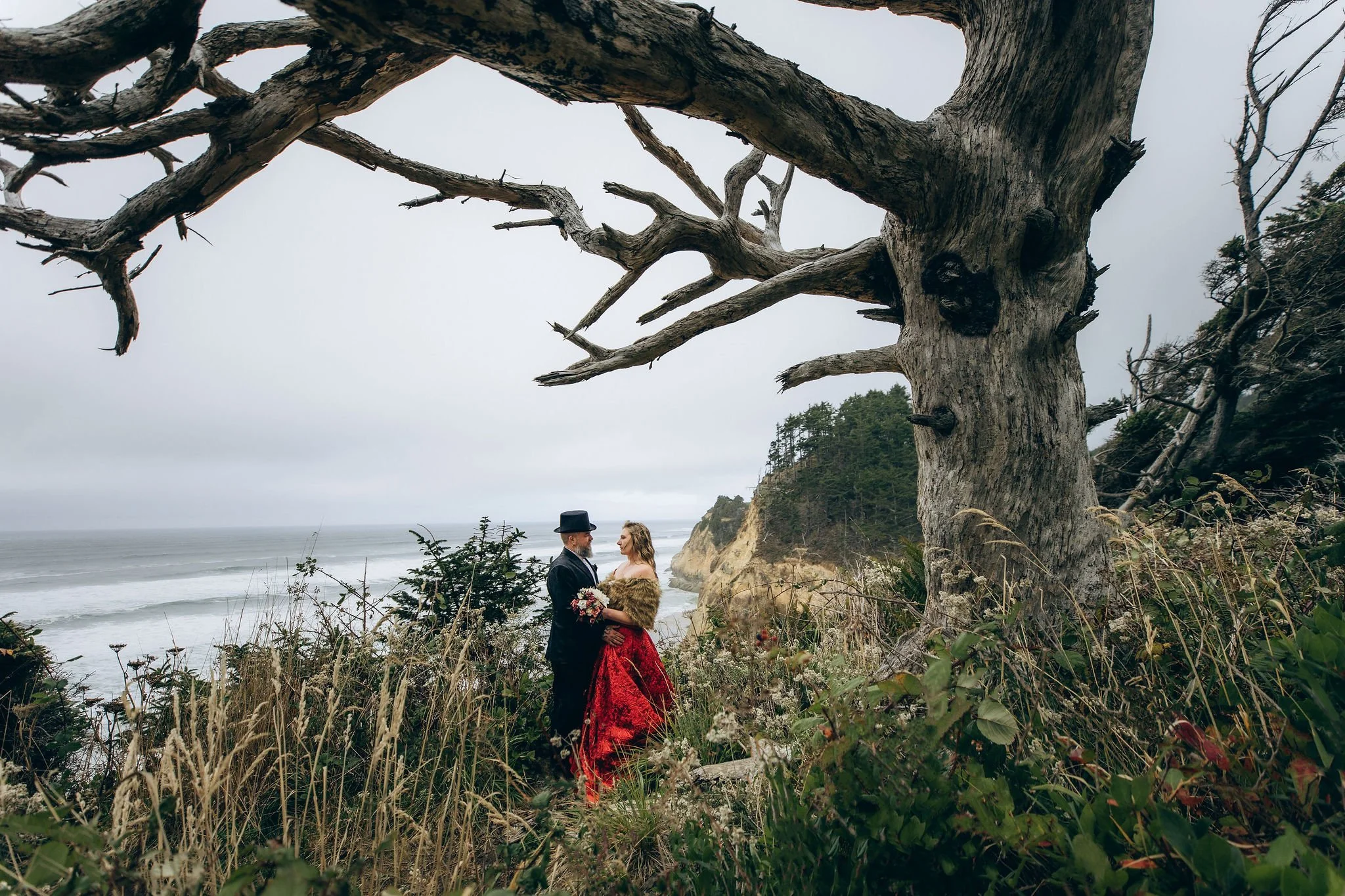 Couple standing together during an elopement at Hug Point on the Oregon Coast, framed by a dramatic dried tree and coastal cliffs