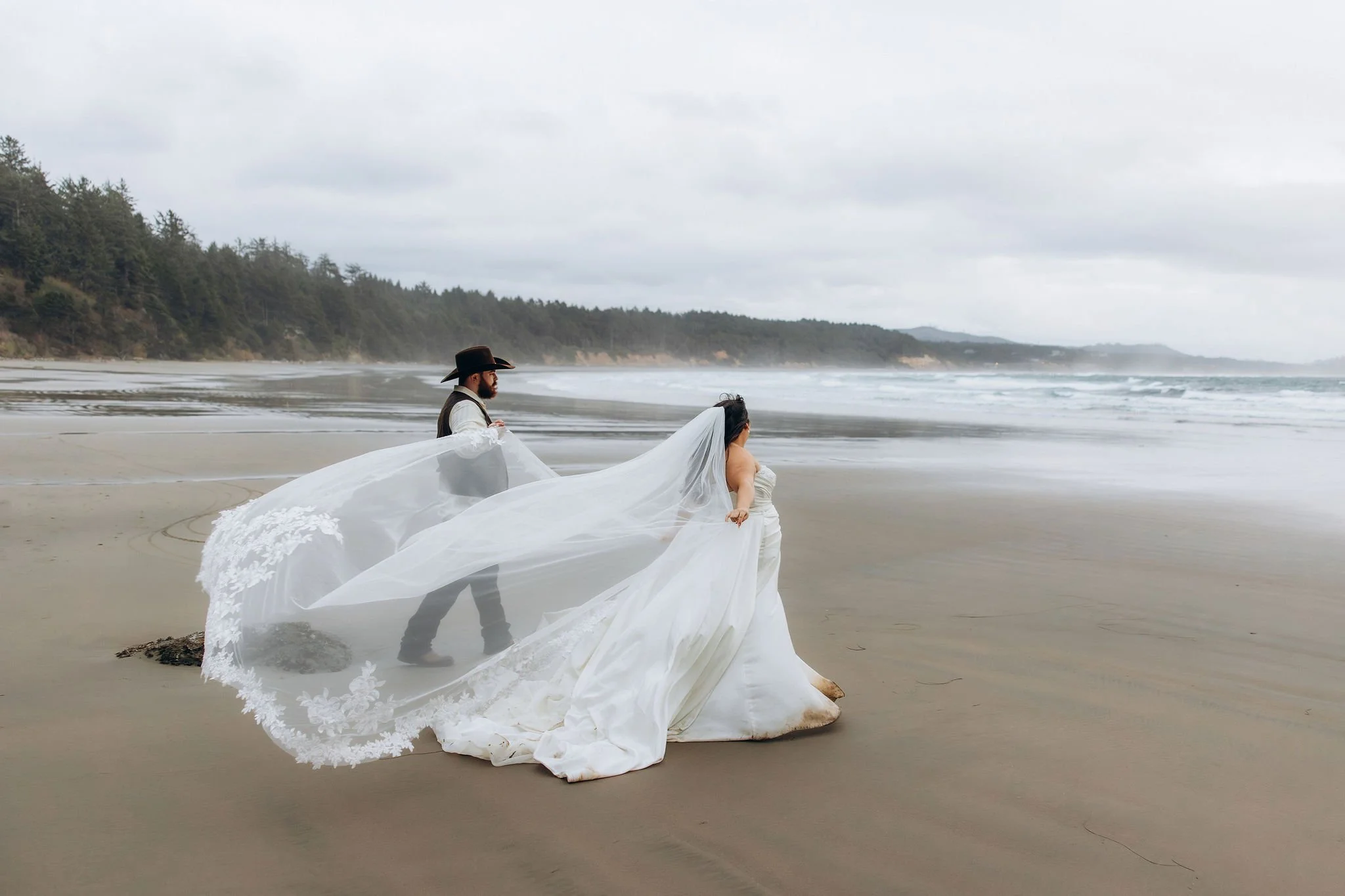 Couple walking along the beach during a sunset elopement at Otter Rock on the Oregon Coast with flowing veil