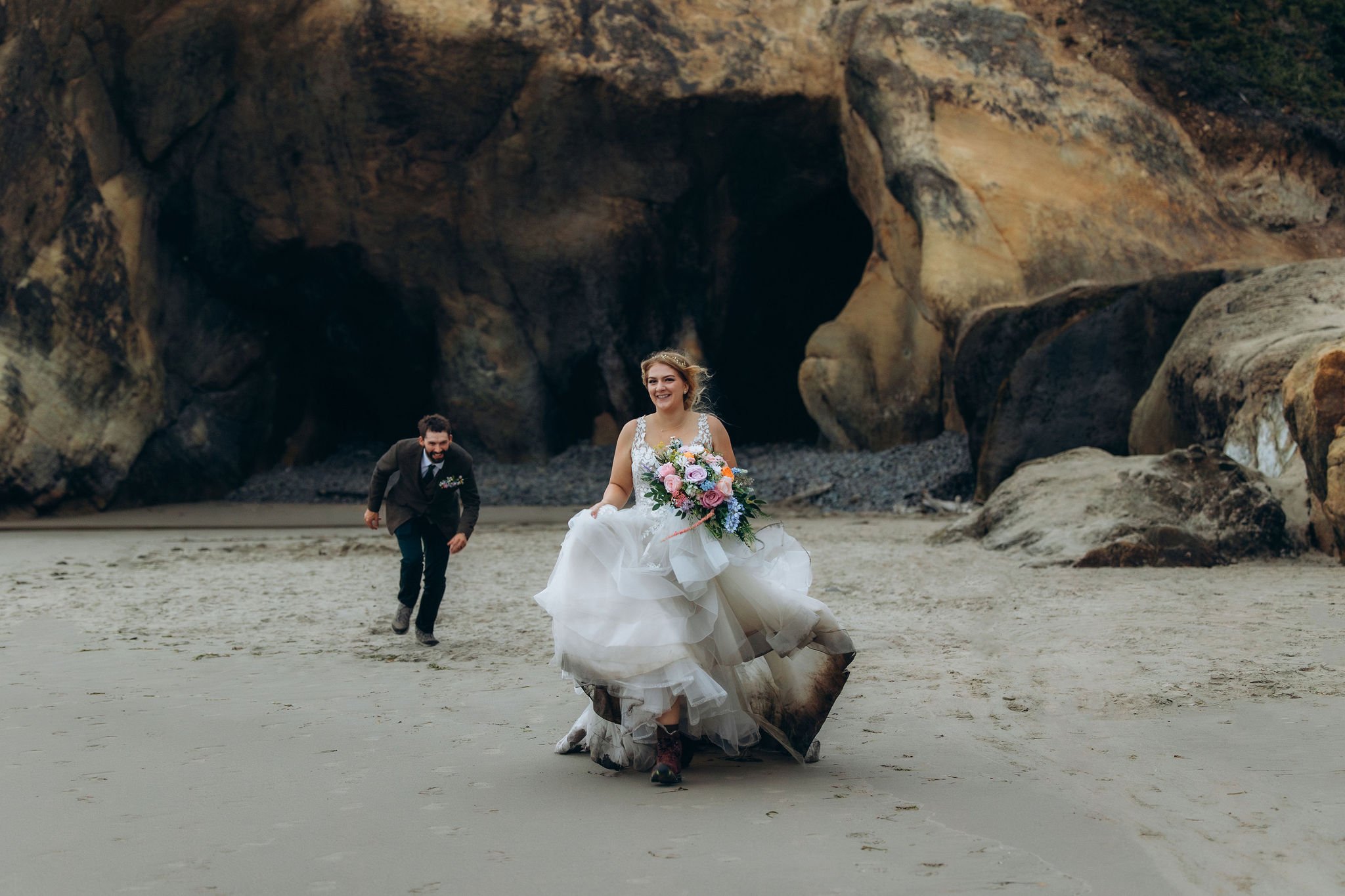 Bride running across the beach at Hug Point on the Oregon Coast during a playful elopement, with rocky cliffs and sea caves in the background