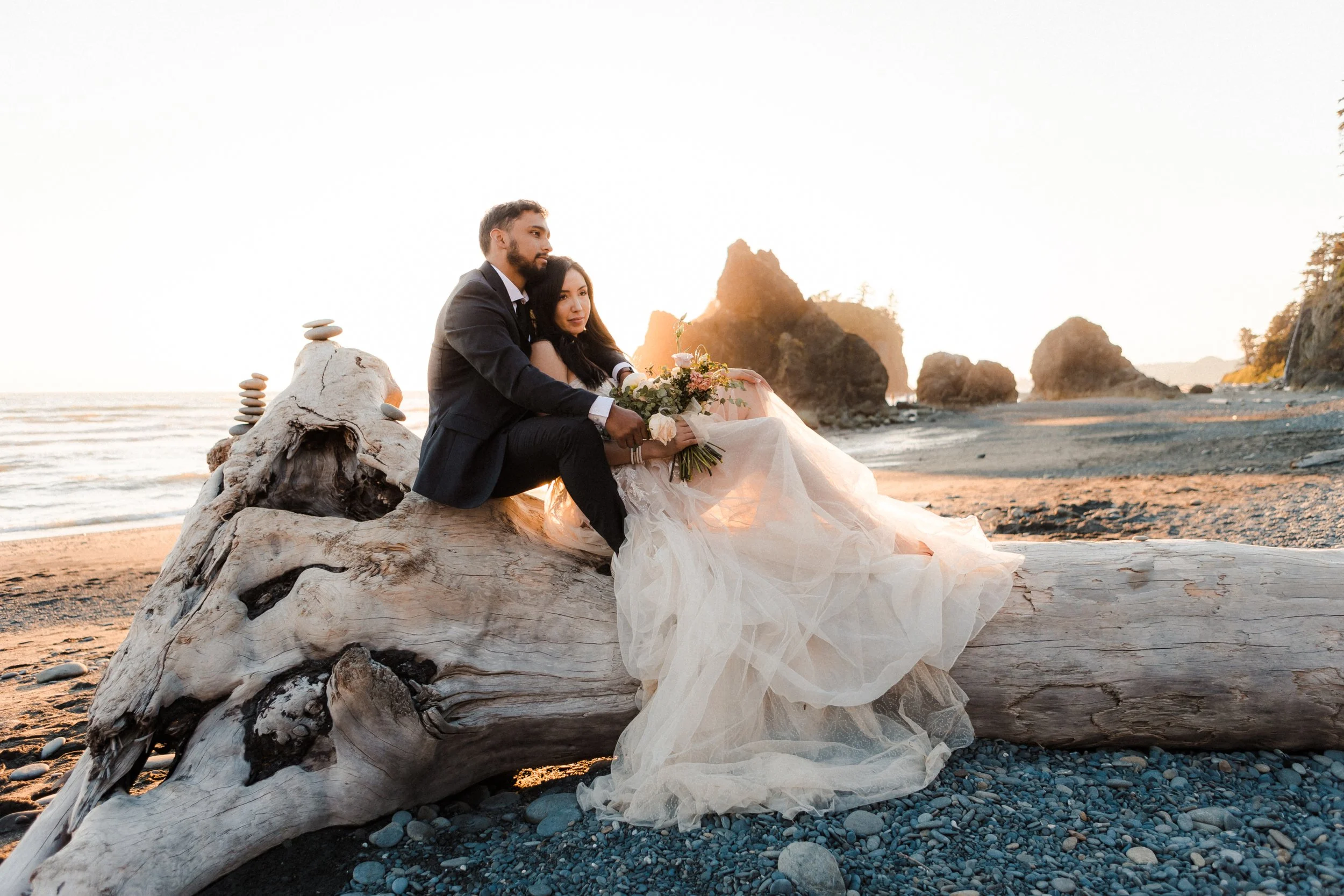 Couple sitting on driftwood during a Pacific Northwest beach elopement at sunset, with the bride wearing a flowy tulle elopement dress that moves beautifully in the ocean breeze