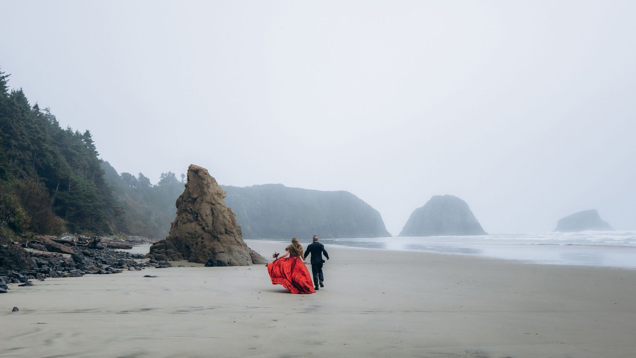 Couple running along Crescent Beach in Ecola State Park, Oregon, during a moody coastal elopement with sea stacks and cliffs in the background