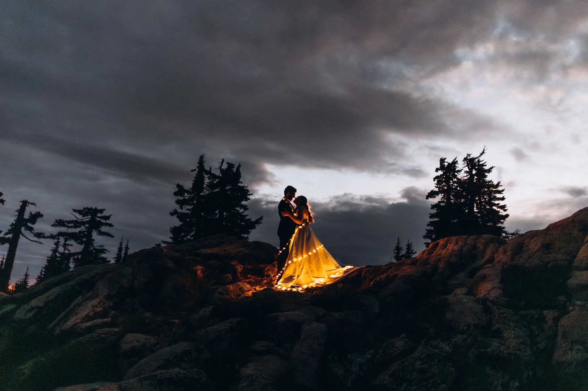 Romantic night mountain elopement with couple standing on rocky overlook, bride wrapped in warm string lights under dramatic cloudy sky
