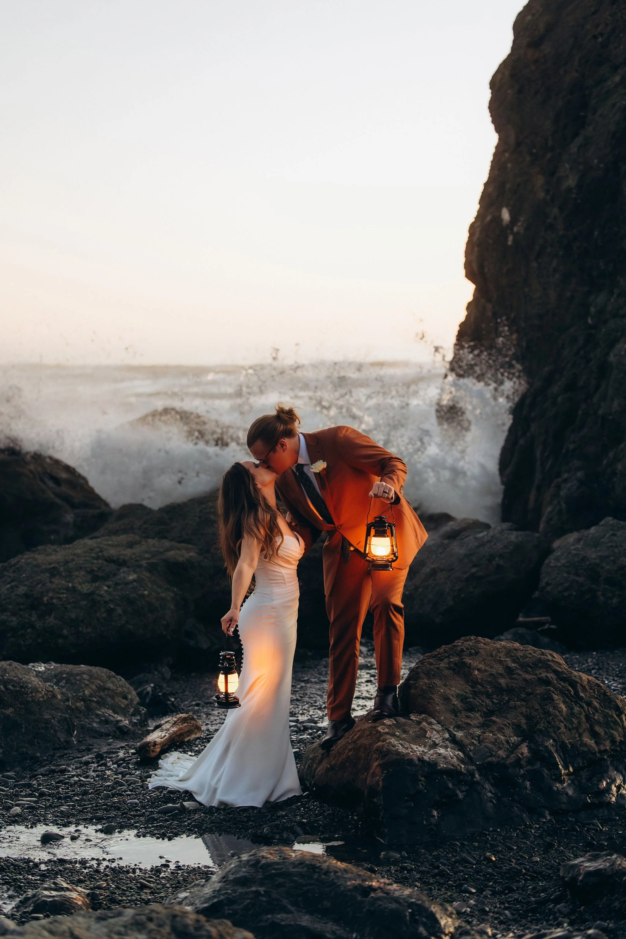 Couple kissing on a rocky beach during sunset elopement in Olympic National Park with crashing waves and lantern light