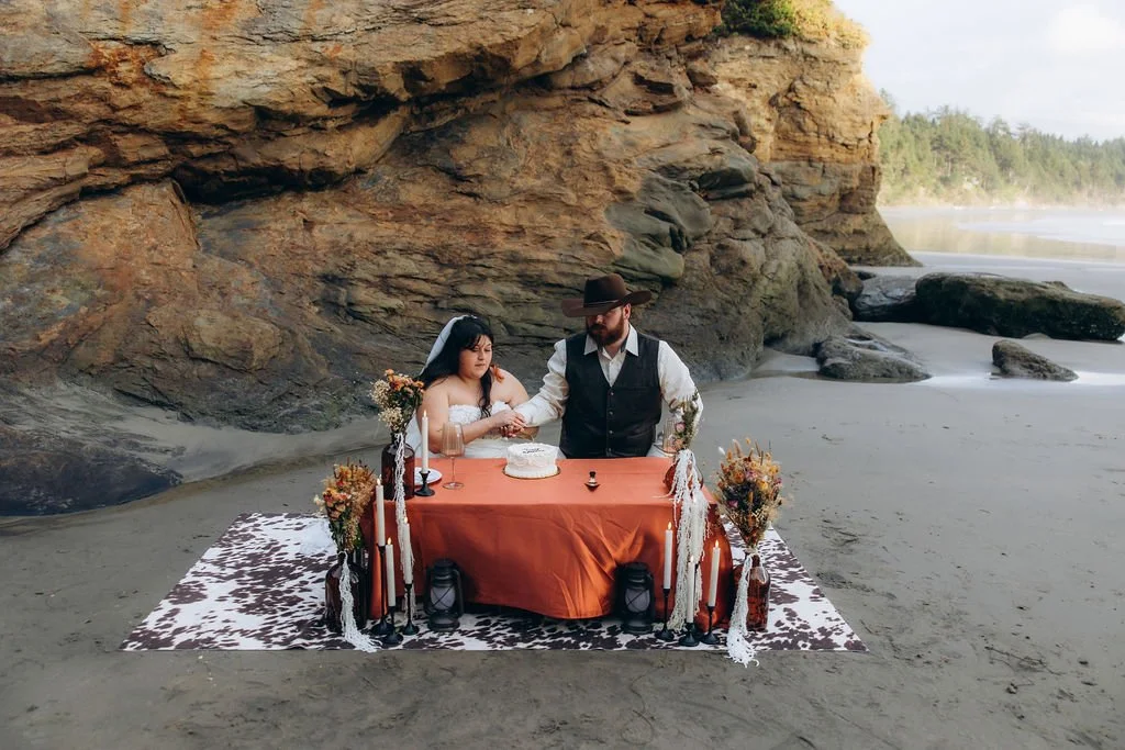 Couple enjoying a picnic celebration during their elopement at Devil’s Punchbowl Beach on the Oregon Coast, seated at a styled table beneath coastal cliffs