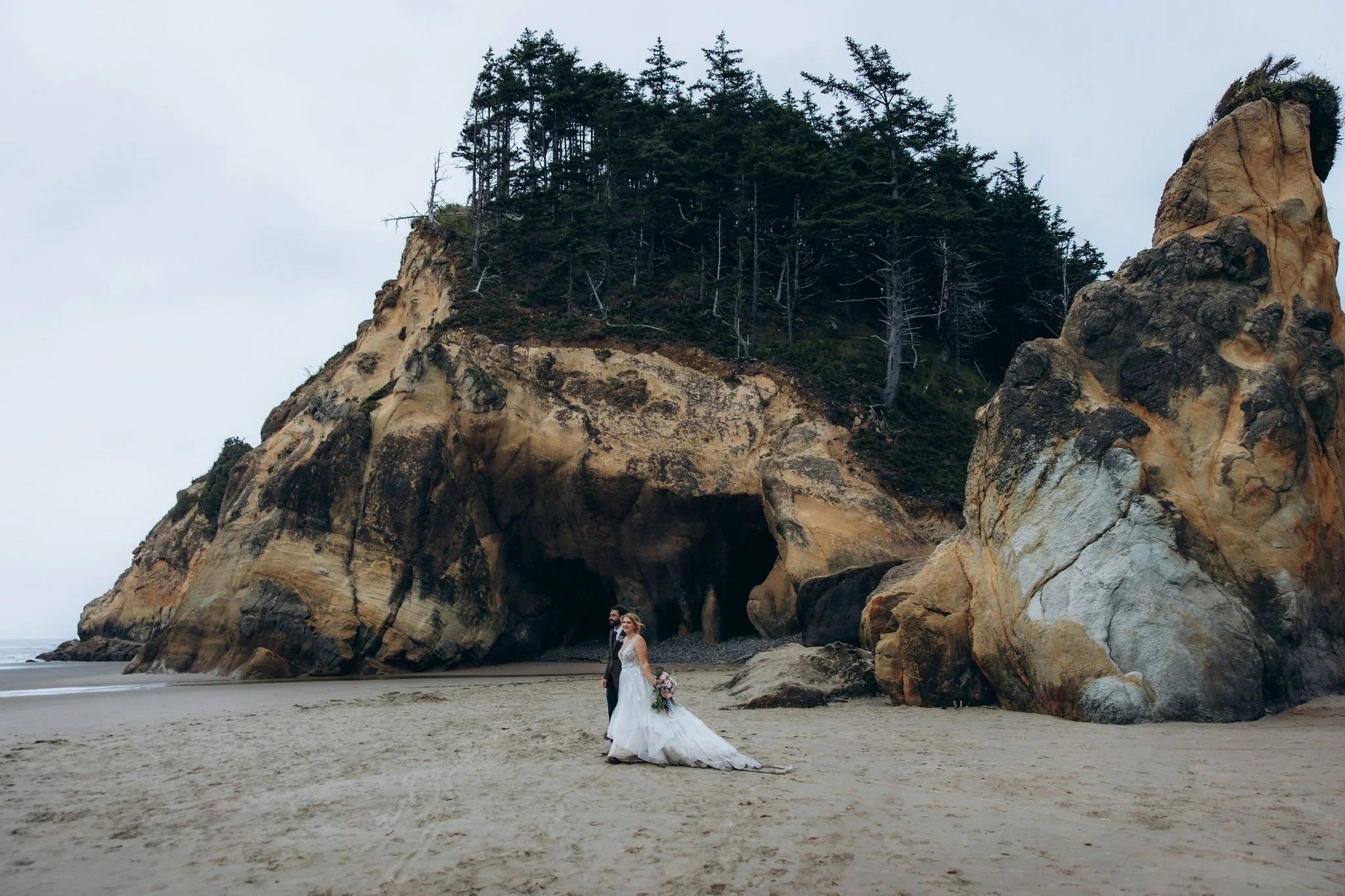 Couple walking along the beach during a sunset elopement at Hug Point caves in Oregon, with sea caves and cliffs visible at low tide