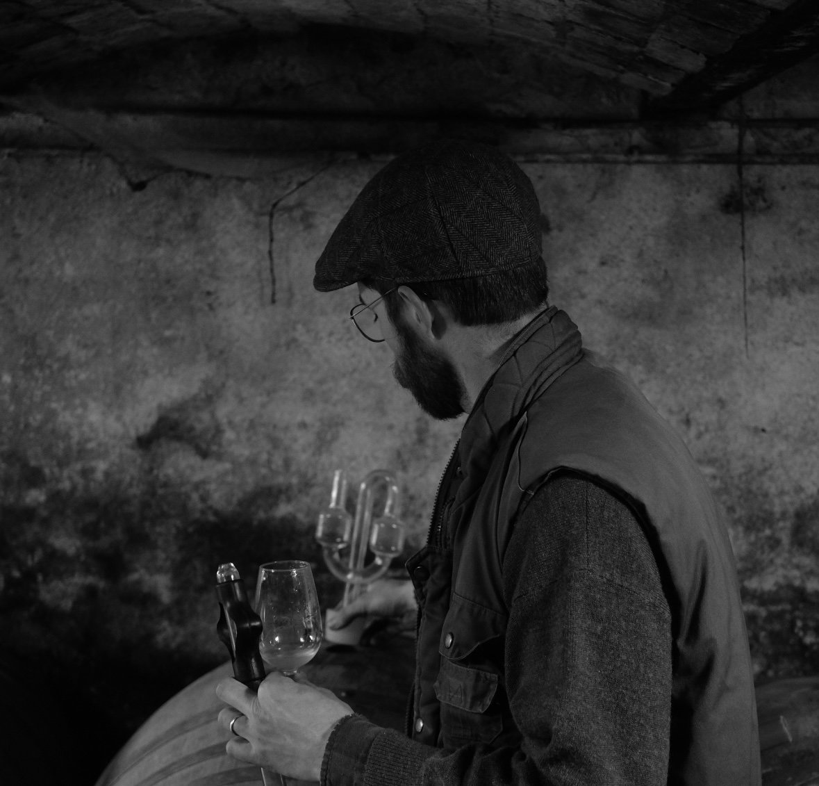 Un homme avec un chapeau, lunettes et barbe, regarde une bouteille de vin et un verre tout en tenant un appareil photo dans une pièce sombre avec un mur en pierre en arrière-plan.