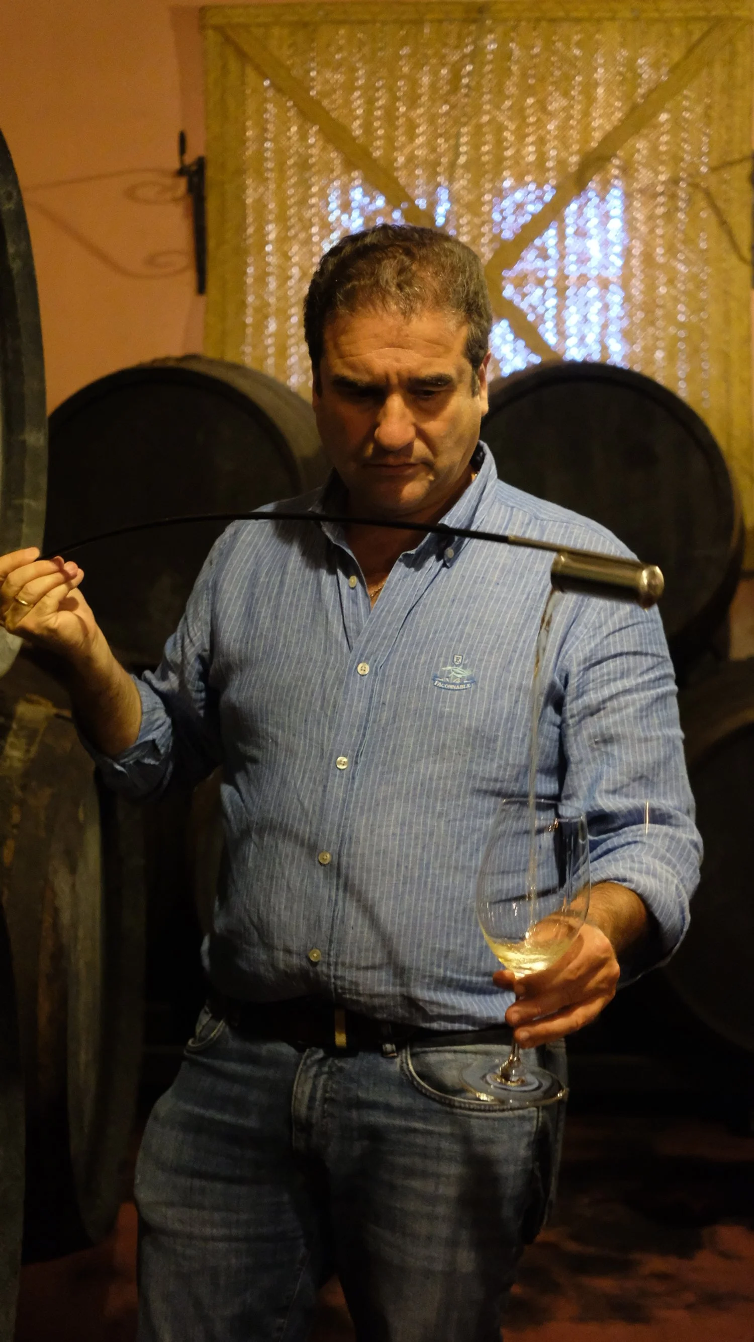 Un homme tient un verre de vin blanc dans une cave à vin et regarde pensif en tenant un tire-bouchon