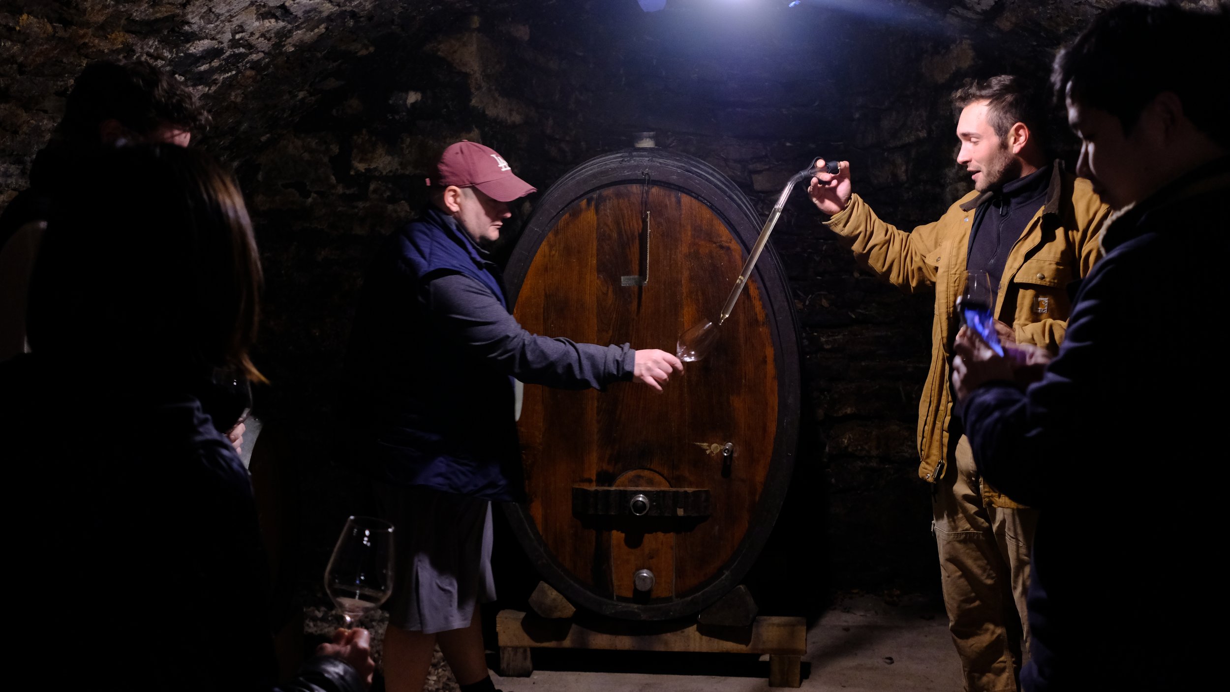 Groupe de personnes dans une cave à vin, certains tiennent des verres de vin et d'autres regardent un homme remplir un verre à partir d'un tuyau qui sort d'une grande cuve en bois.
