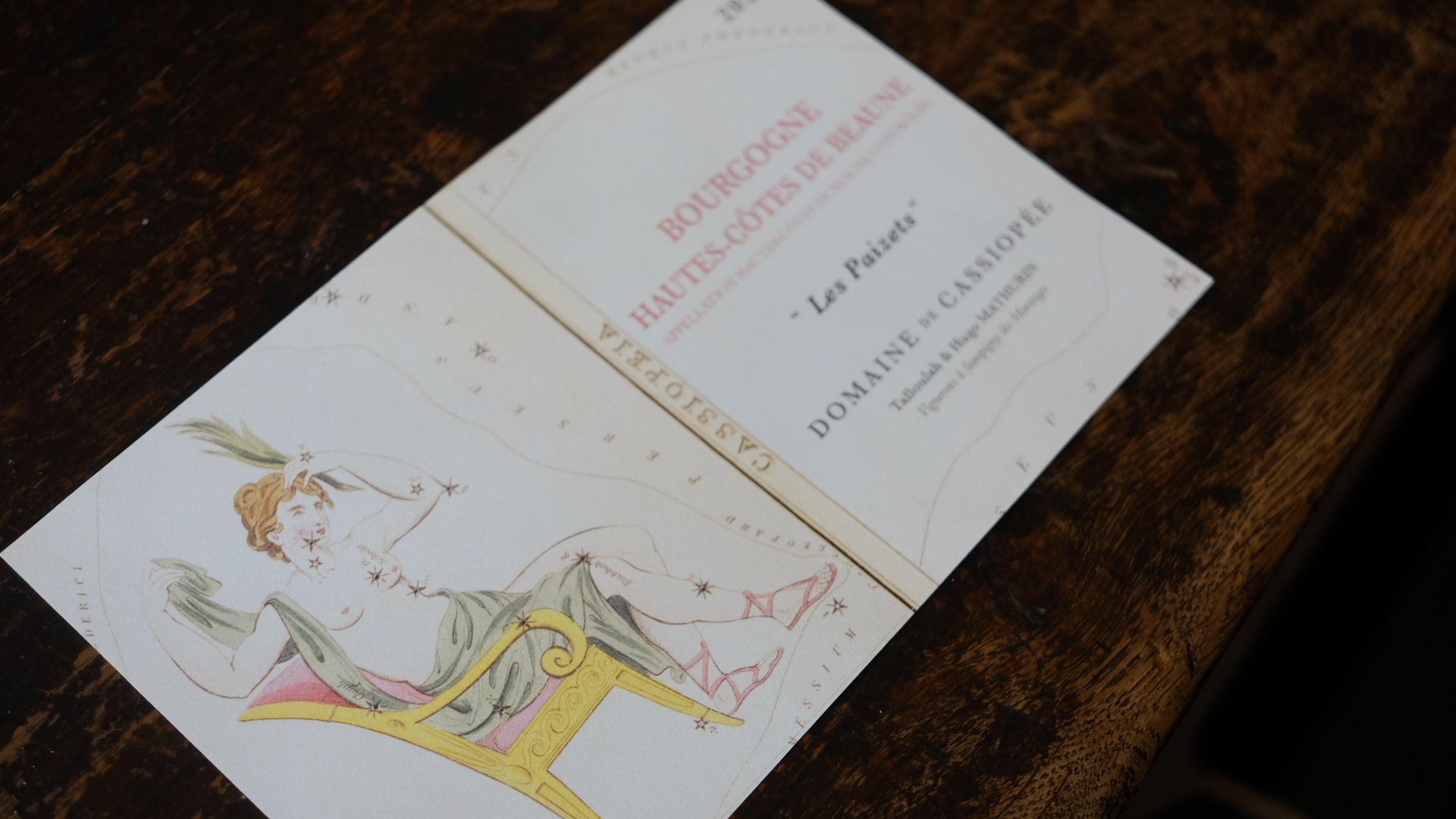 Une brochure de vin posée sur une table en bois foncé. La brochure comporte une illustration d'une femme avec une couronne de trèfles et une écharpe verte, assise sur une chaise dorée, tenant un trèfle dans sa main. Le texte de la brochure est en fra