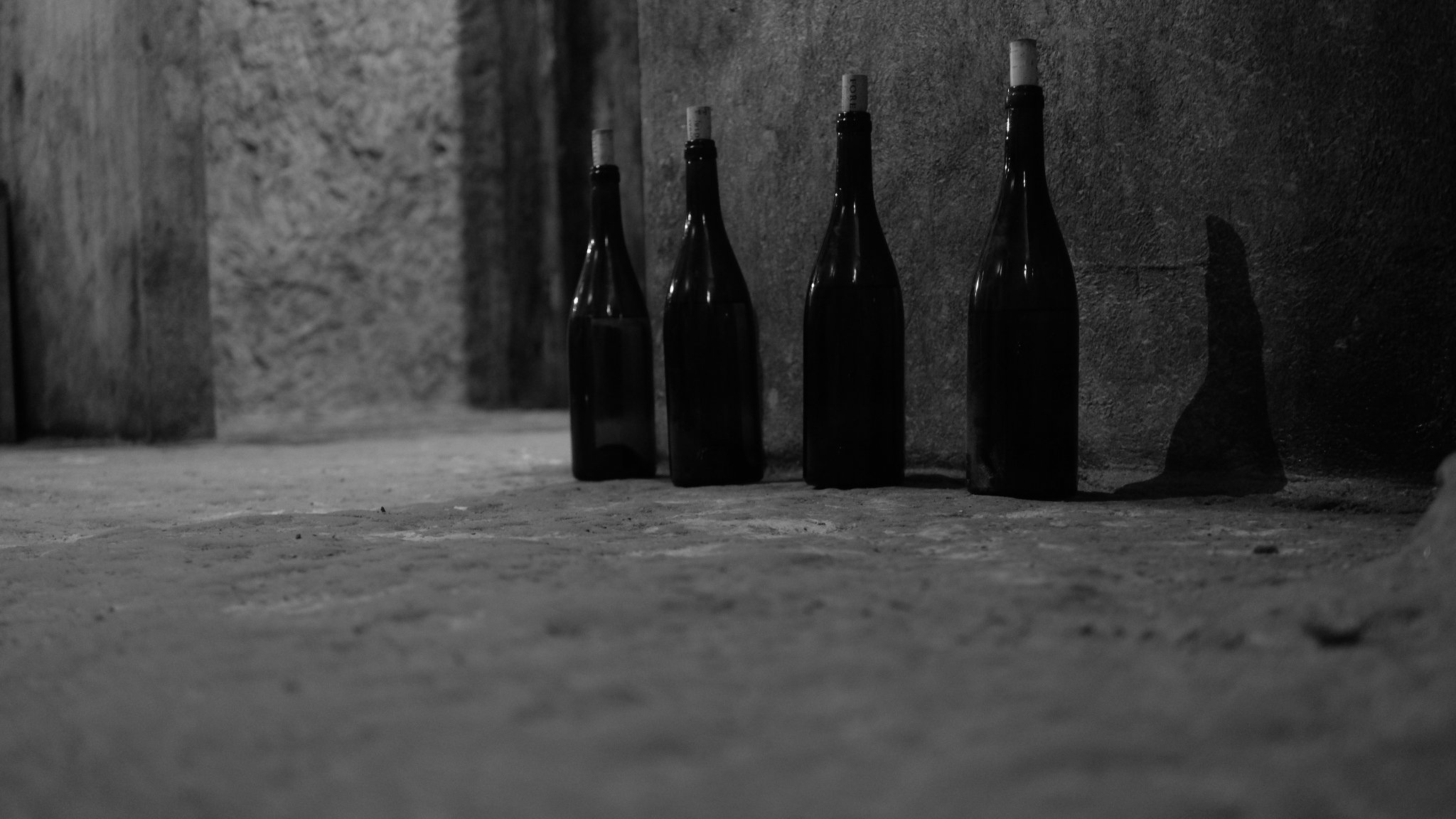 Quatre bouteilles de vin en verre sombre alignées sur un sol en pierre dans une cave sombre, avec un mur en pierre en arrière-plan.