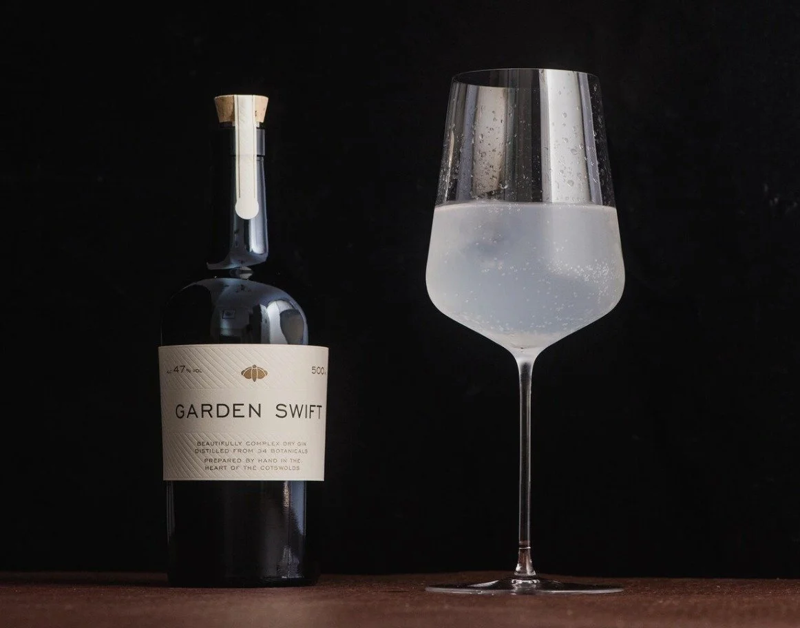 Une bouteille de gin nommée GARDEN SWIFT à côté d'un verre de vin blanc pétillant sur une surface en bois avec un fond noir.
