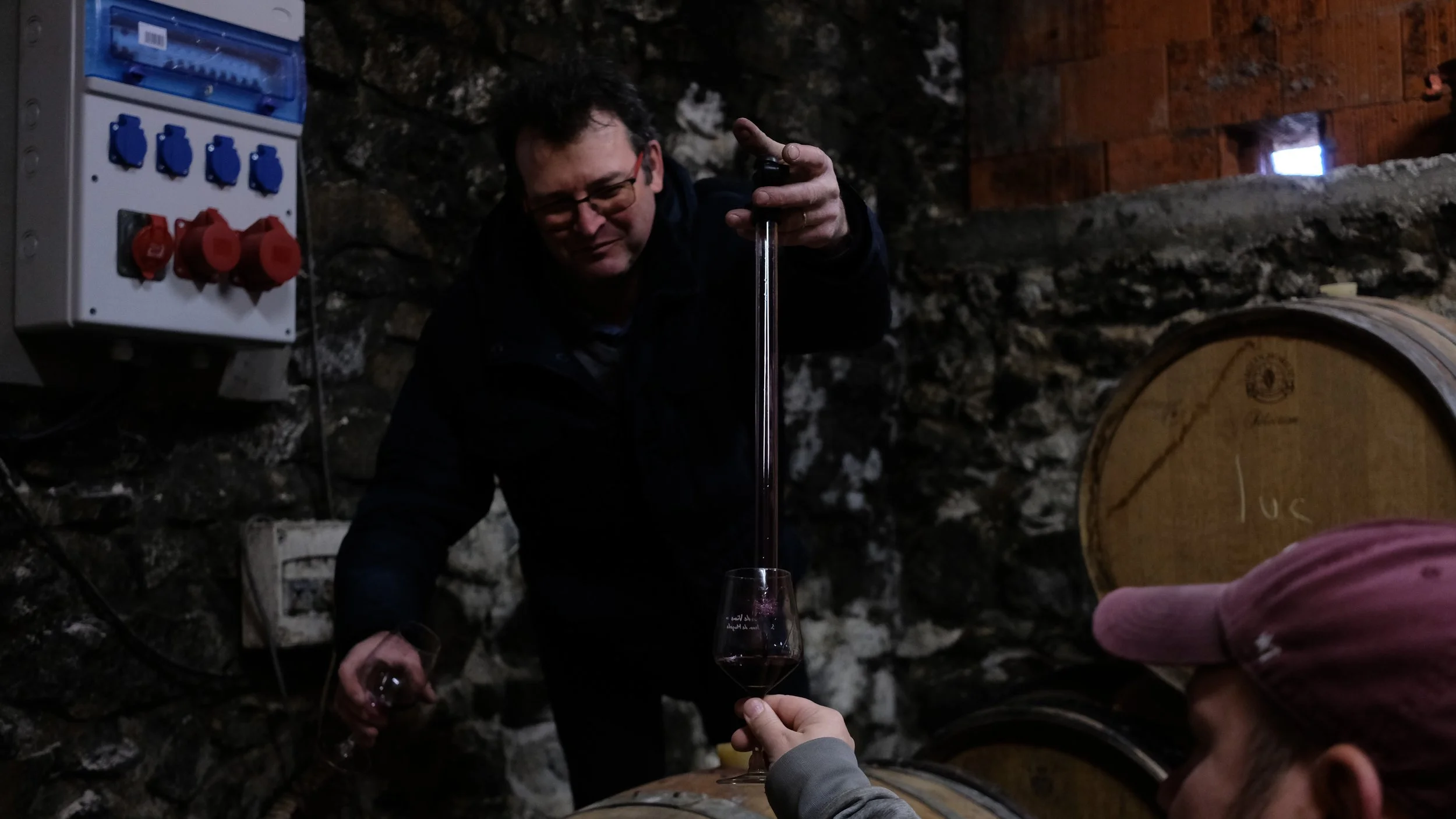 Une personne verse du vin dans un verre depuis un tube en burette dans une cave à vin, avec des cuves en bois en arrière-plan.