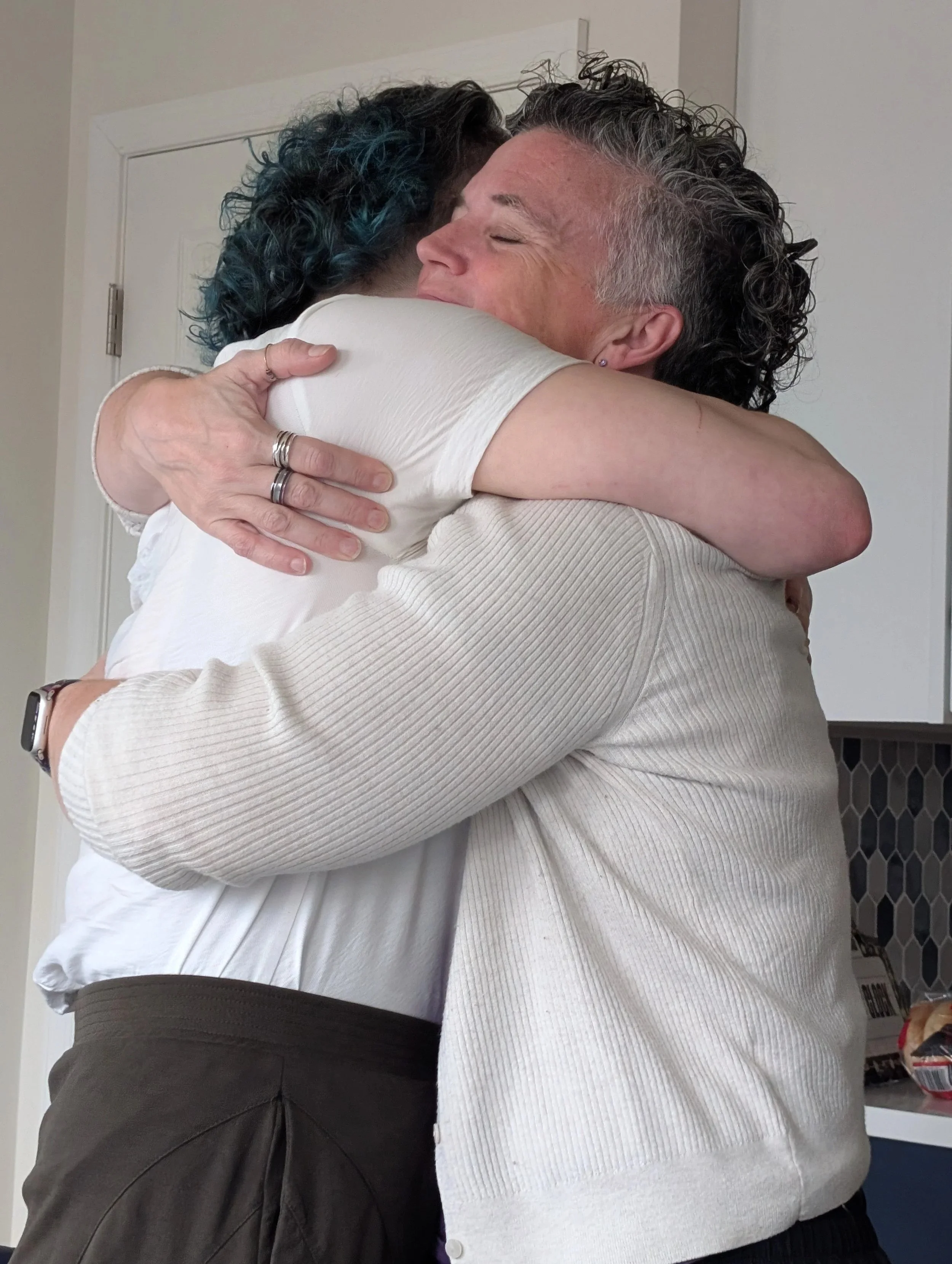 Two individuals embracing, one with short dark hair and the other  (Michelle Renee) with curly gray hair, in a warm hug indoors.