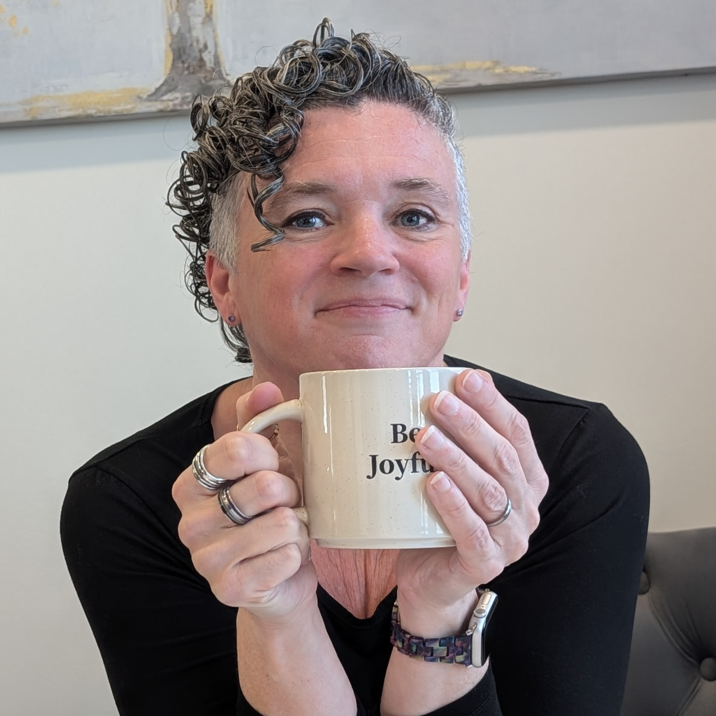 A woman (Michelle Renee)  with short curly gray hair, holding a coffee mug with the words "Be Joyful" printed on it, wearing a black top and jewelry, sitting in a room with a painting on the wall.